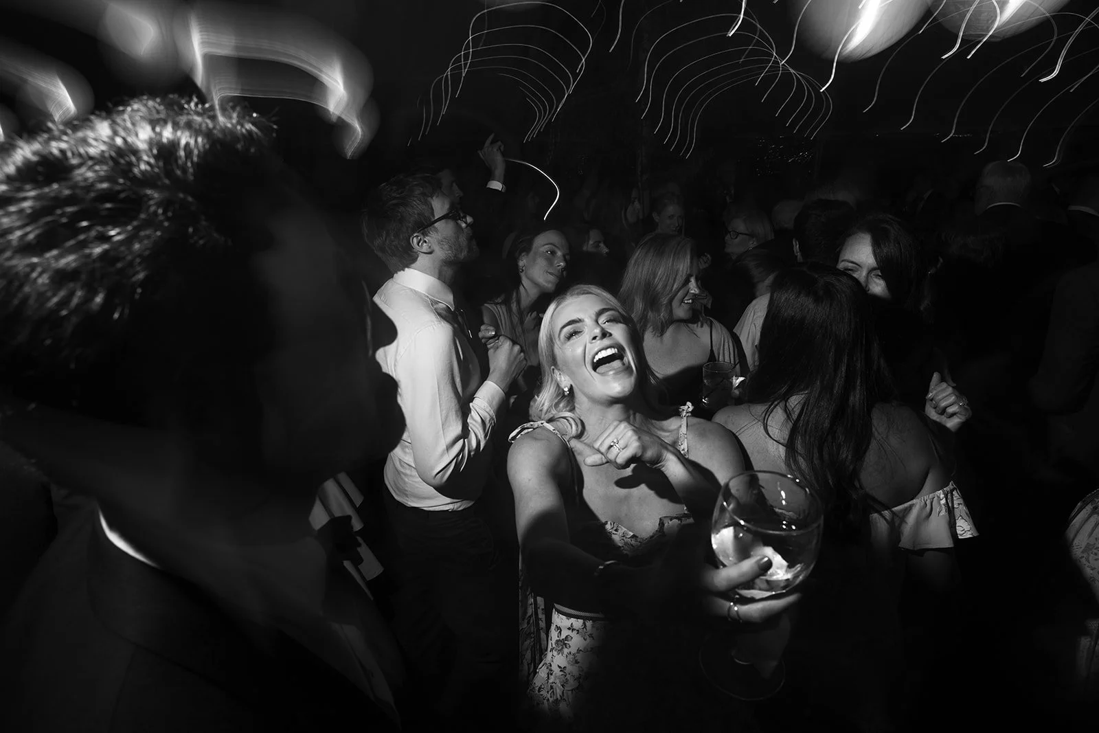 A group of people dancing and socializing at a party, with a woman in the center holding a drink and laughing joyfully.