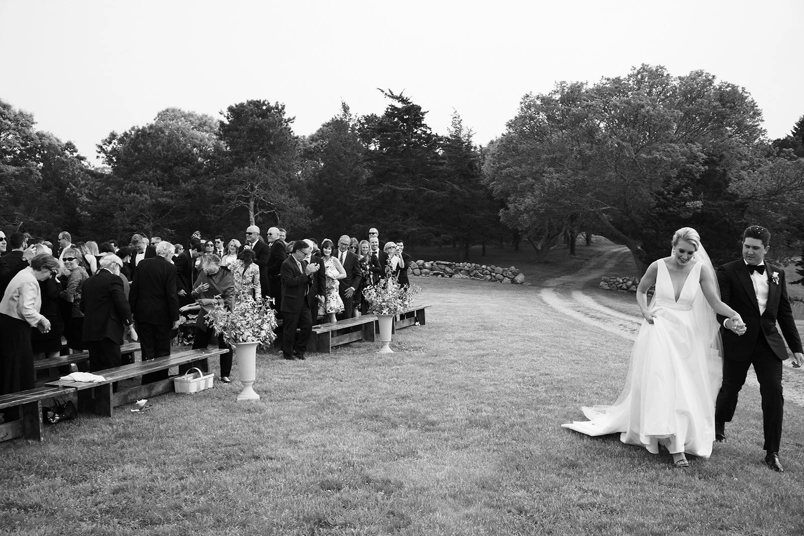 Black and white photo of a wedding celebration outdoors. A bride and groom walk hand in hand on the grass, with guests gathered on benches and standing nearby, watching and taking photos. Trees large and small surround the scene.