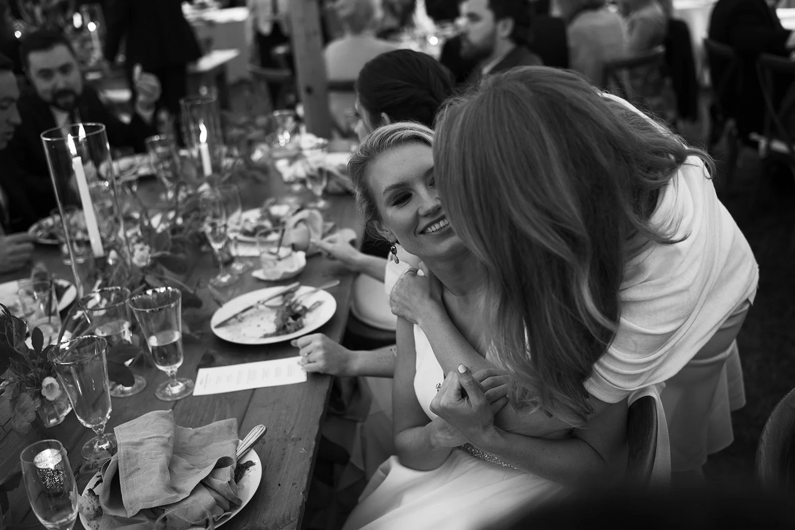A woman in a white dress smiling while being hugged by another woman at a formal dinner party with a table set with glasses, plates, and candles.