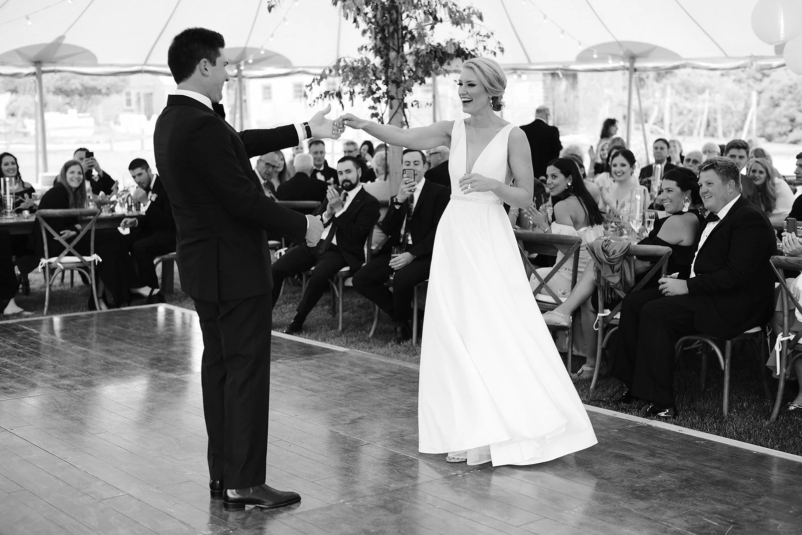 A black and white photo of a wedding reception. A bride and groom are sharing a dance under a large tent with guests seated and watching, some taking photos and clapping.