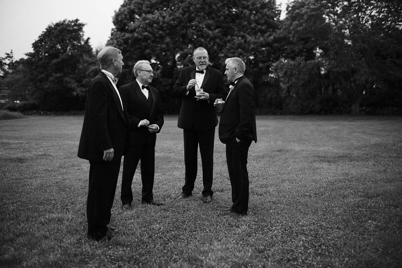 Five men dressed in formal tuxedos standing on a grassy field and engaging in conversation, with trees in the background, in black and white.
