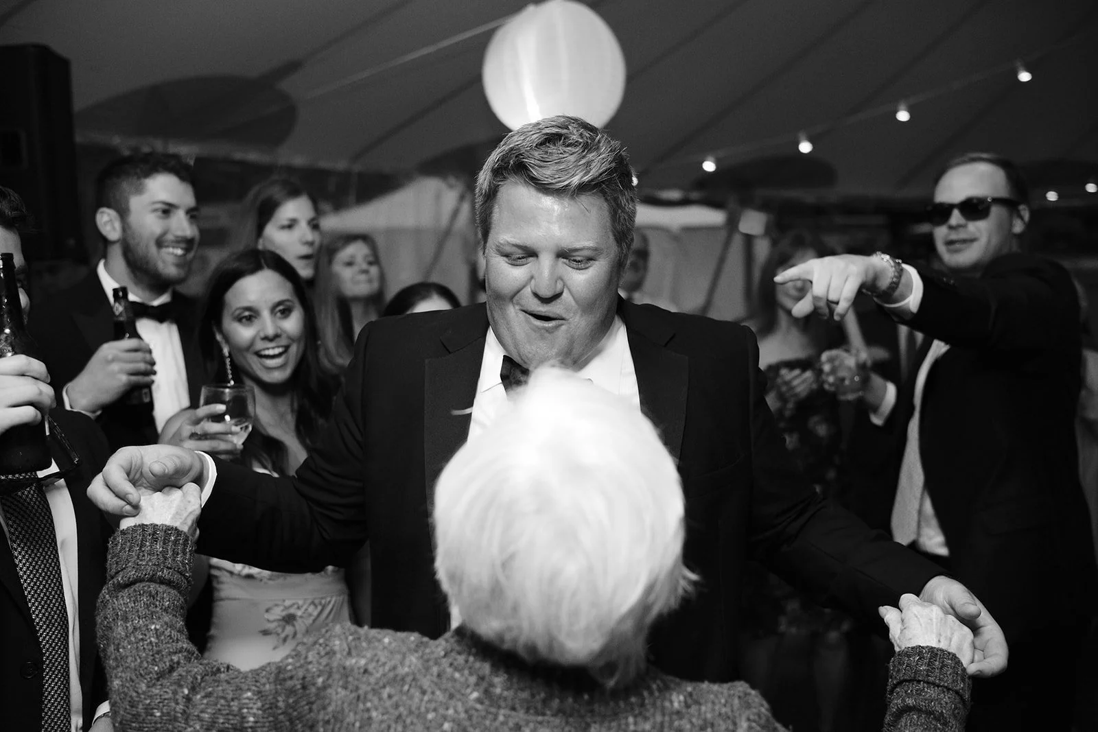 People dancing and socializing at a party, dressed in formal attire, with a man in a tuxedo holding hands with an elderly woman, in a large decorated event tent