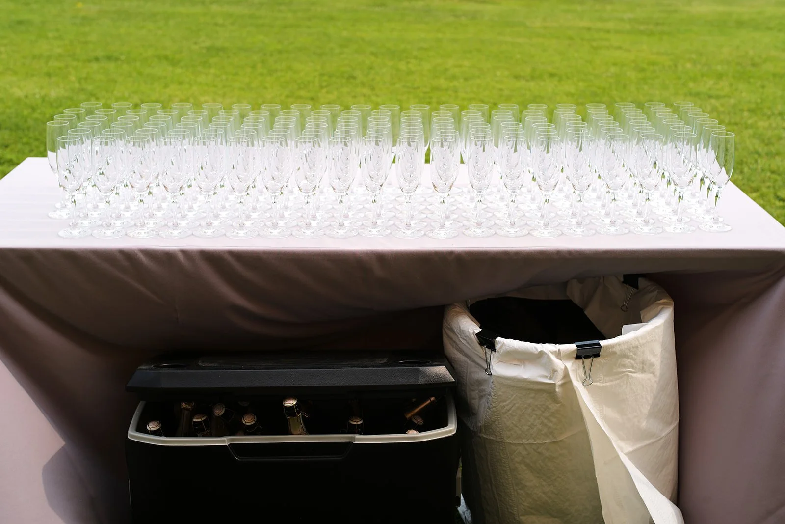 A table with numerous empty champagne flutes set on it, with containers and a grassy outdoor space in the background.