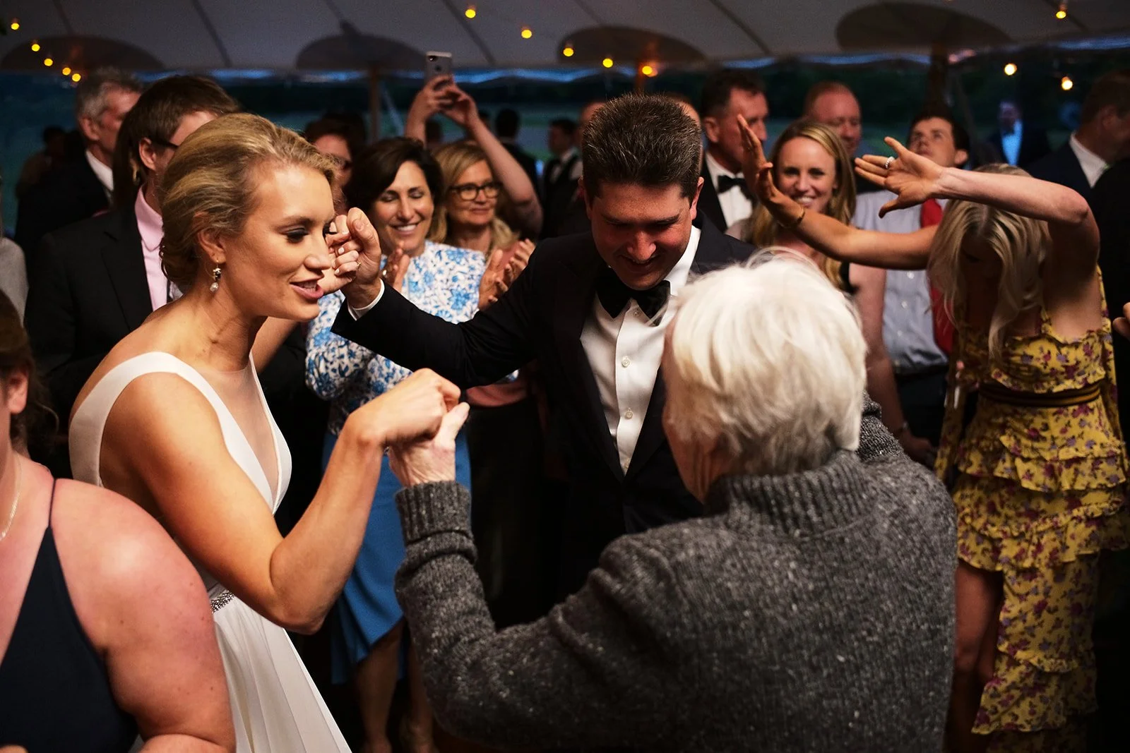 Crowd of people dancing and celebrating at a wedding reception, with a couple, possibly the bride and groom, in the center dancing and smiling, surrounded by guests clapping and enjoying the moment.