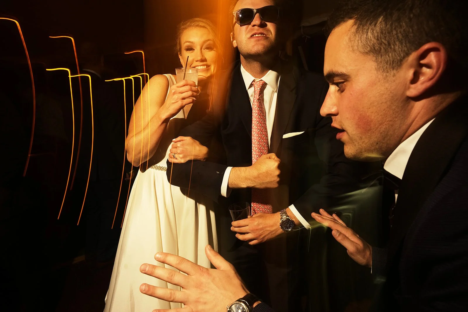 People at a party, dressed in formal attire, with a woman holding a drink and men in suits, in a dimly lit setting.