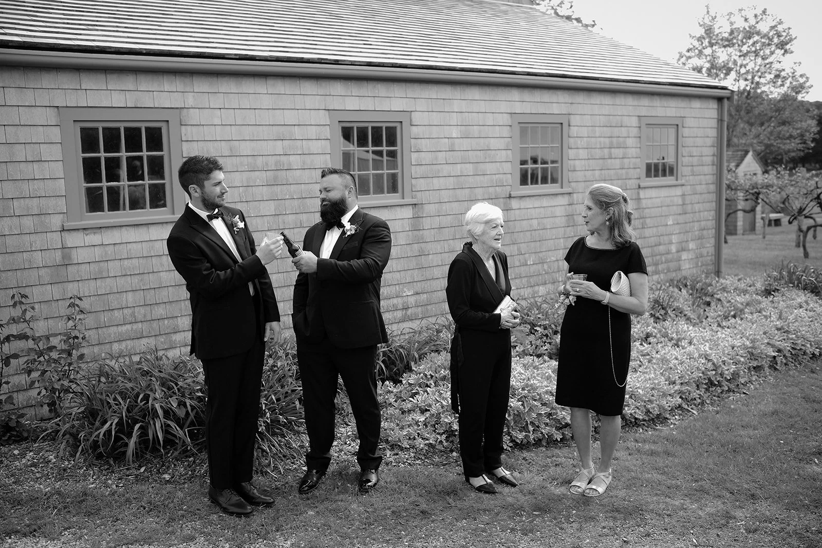 Four people in formal attire standing outside a building with shingled walls, engaged in conversation and holding drinks.