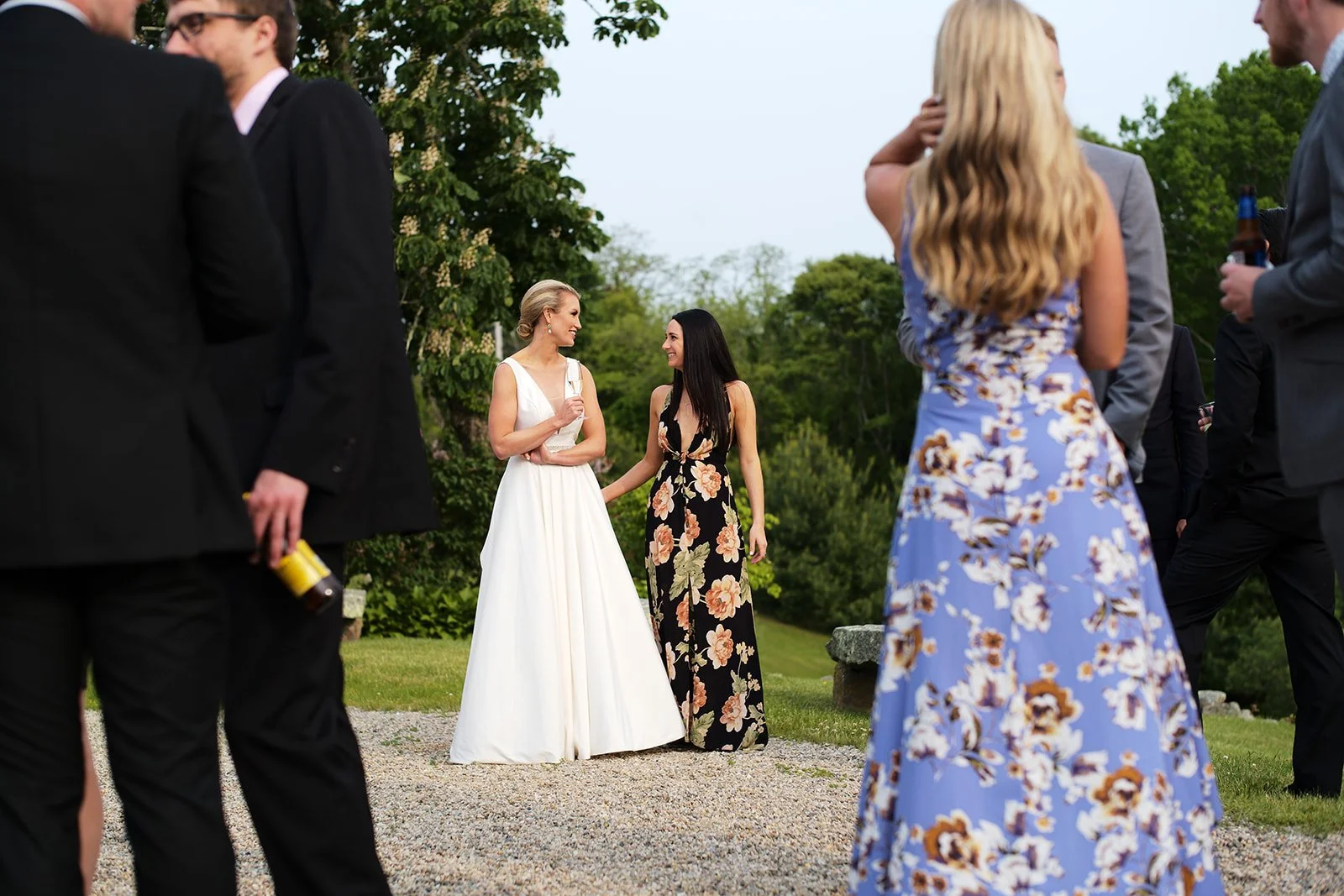 People attending an outdoor social gathering, with two women talking in the background, one in a white dress and the other in a floral dress, while others mingle around in formal attire.