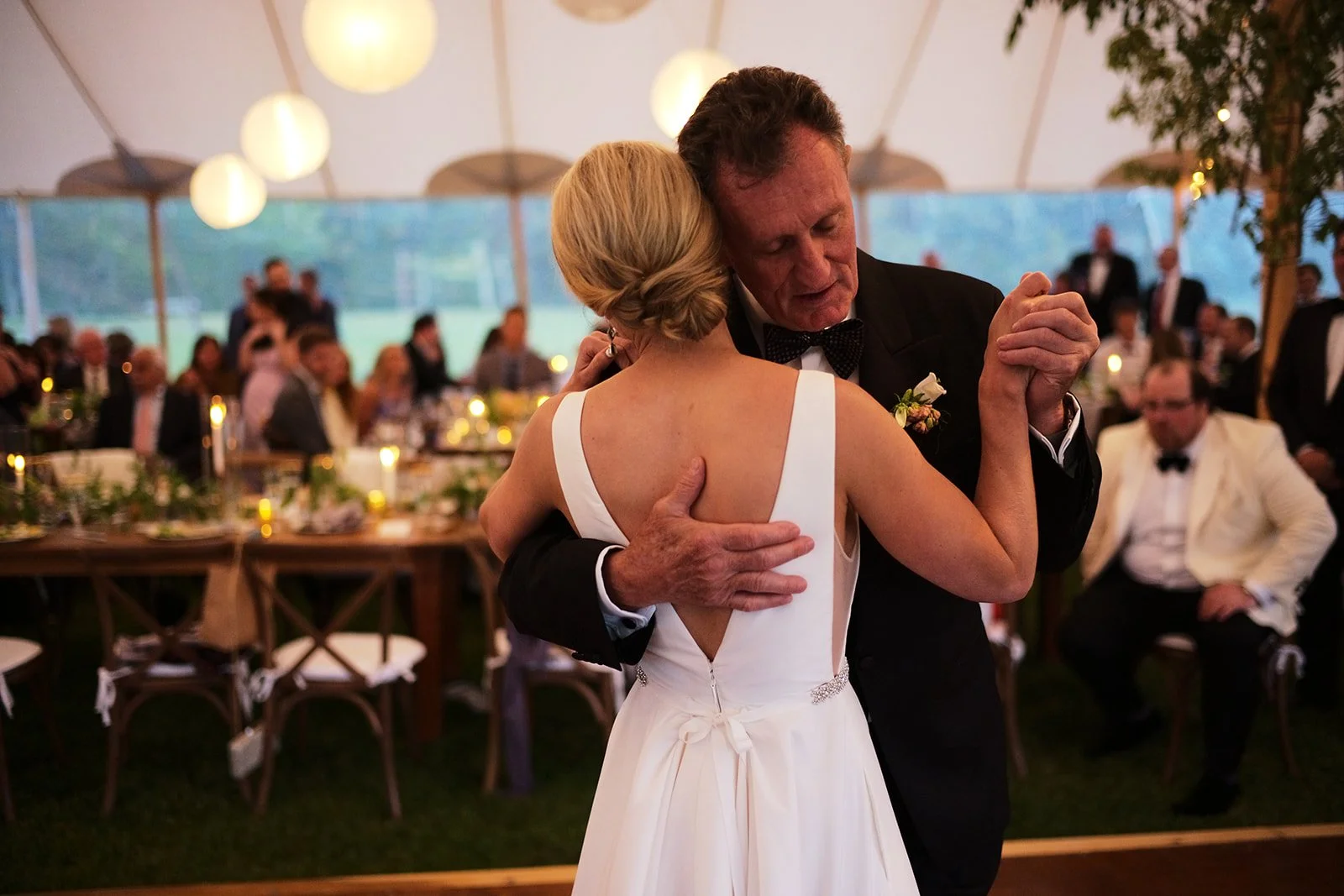 A couple dancing closely at a wedding reception under a large tent, with guests seated at decorated tables in the background.