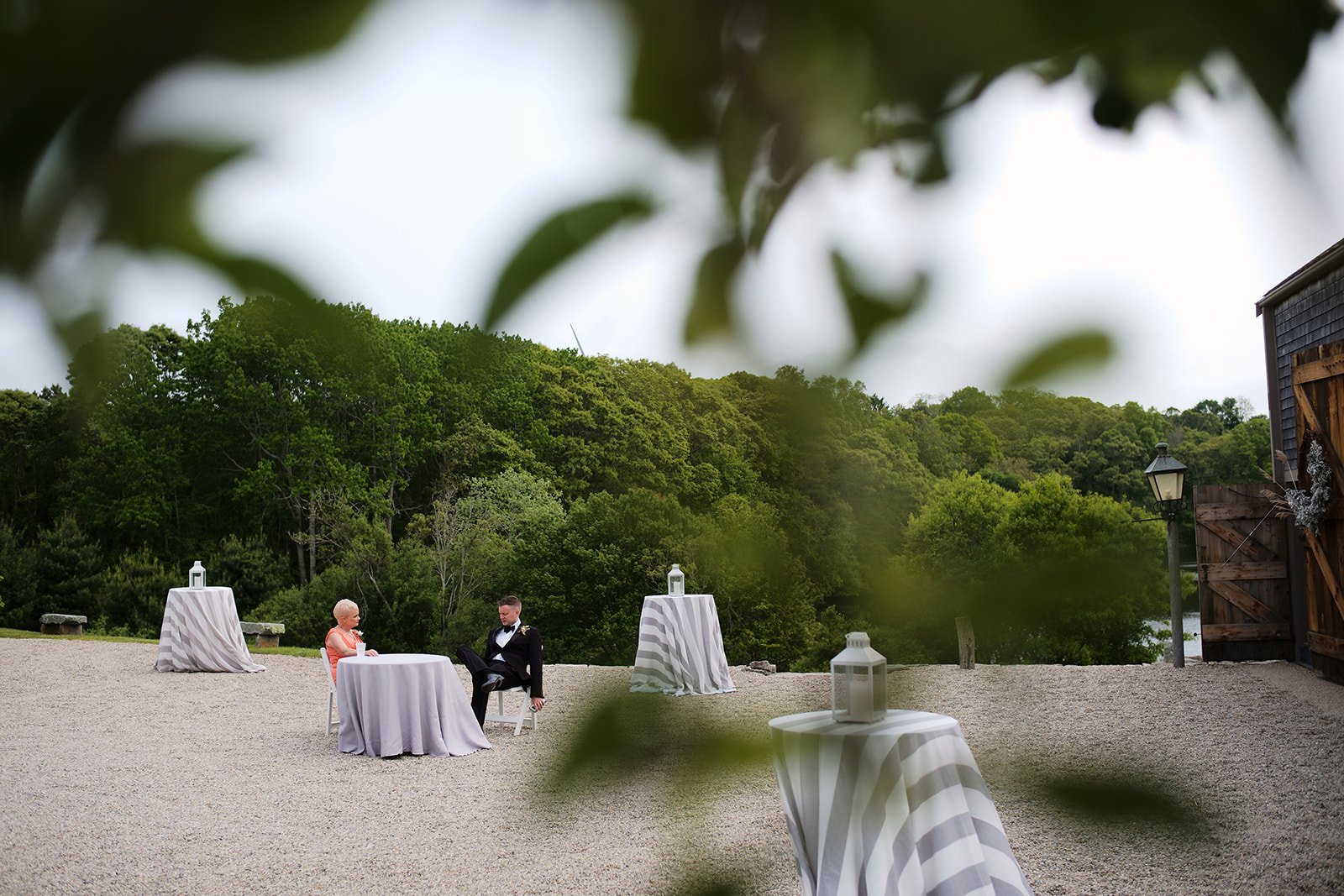 An outdoor wedding reception setup with tables covered in striped tablecloths, lanterns on each table, a couple seated at one table, and a scenic background of green trees and a cloudy sky.