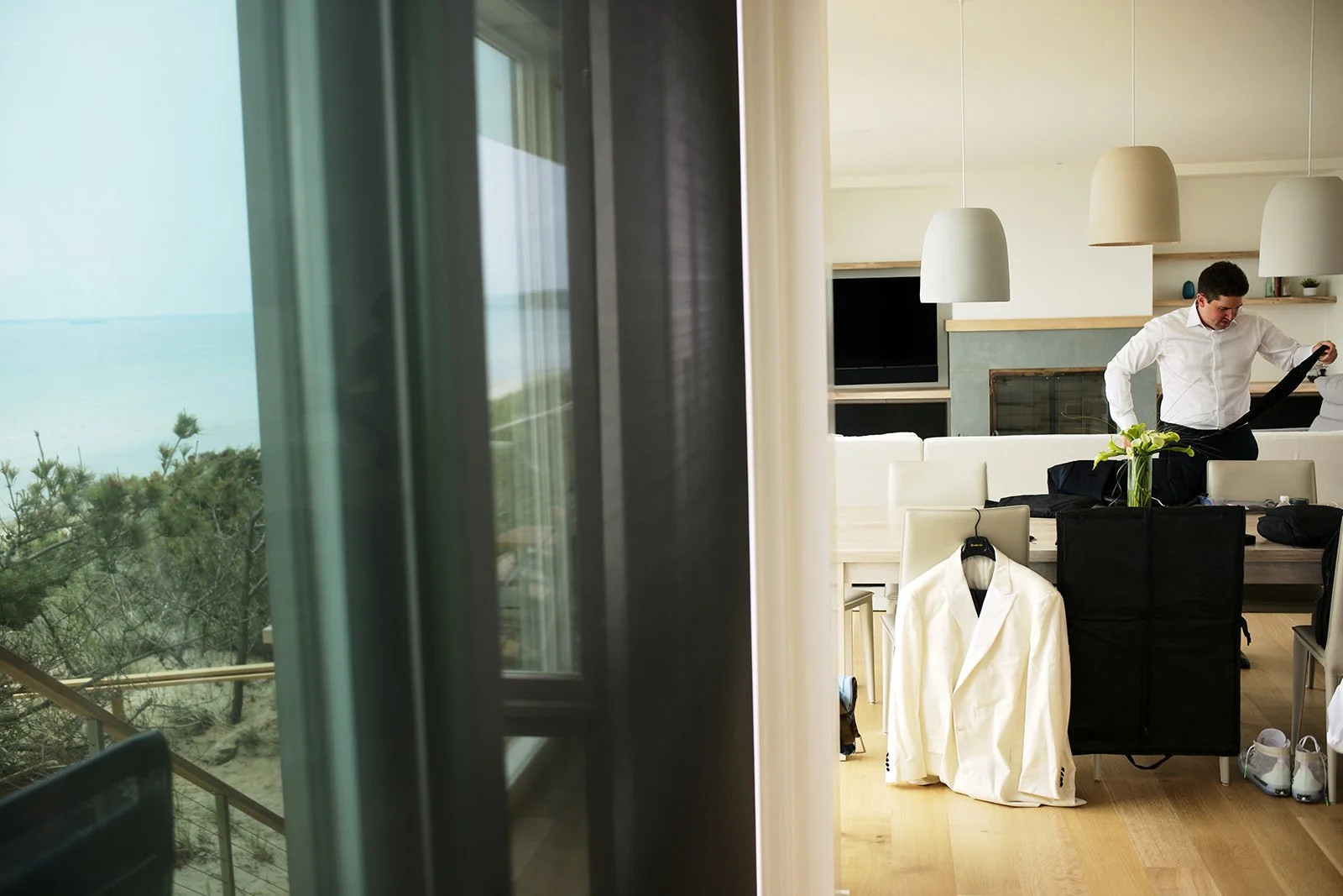 A man in a white shirt preparing for a formal event in a modern, bright living room with a view of trees and a beach outside.