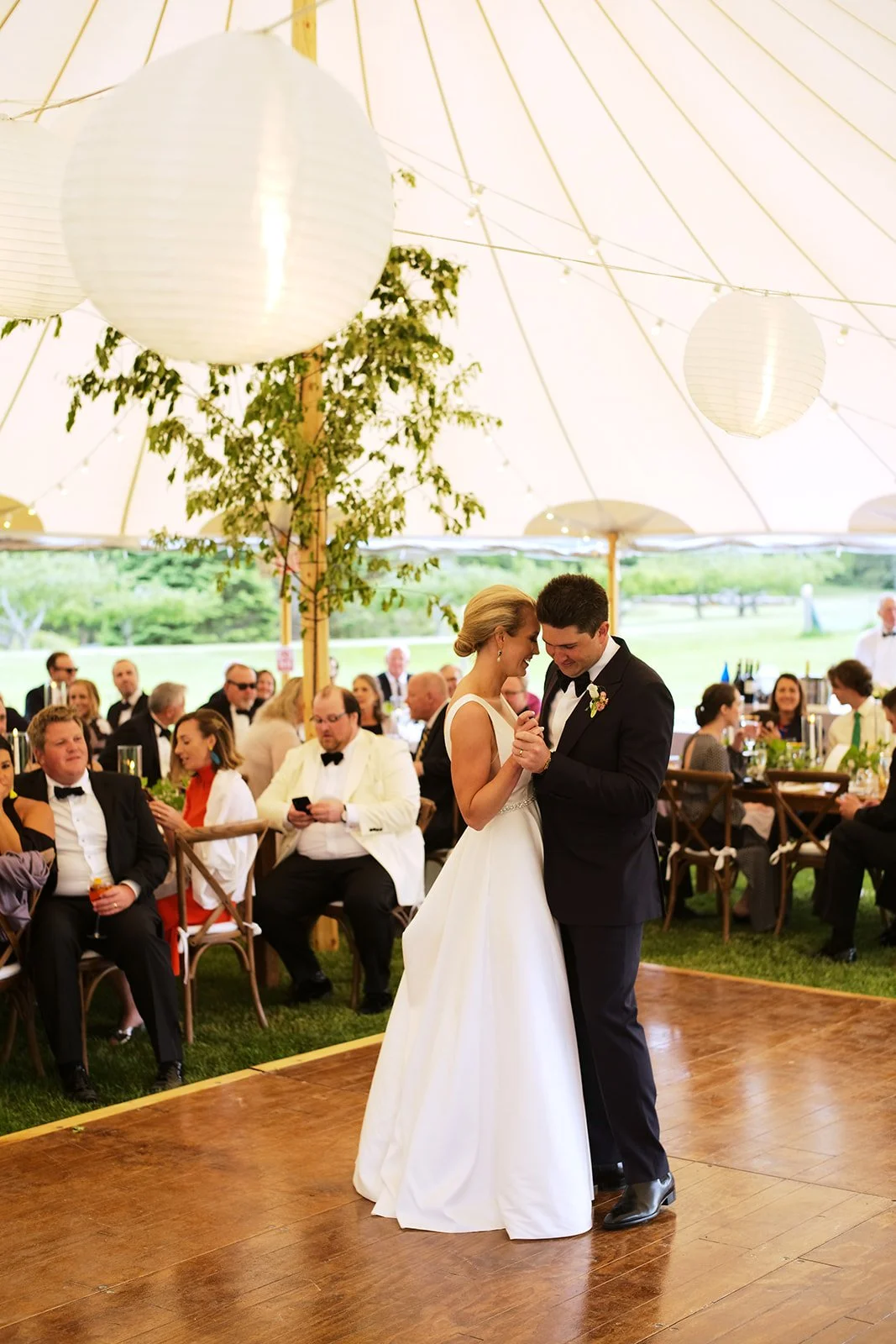 A bride and groom dancing at a wedding reception under a large white tent, with guests seated around them, some holding drinks, and paper lanterns hanging from the tent ceiling.