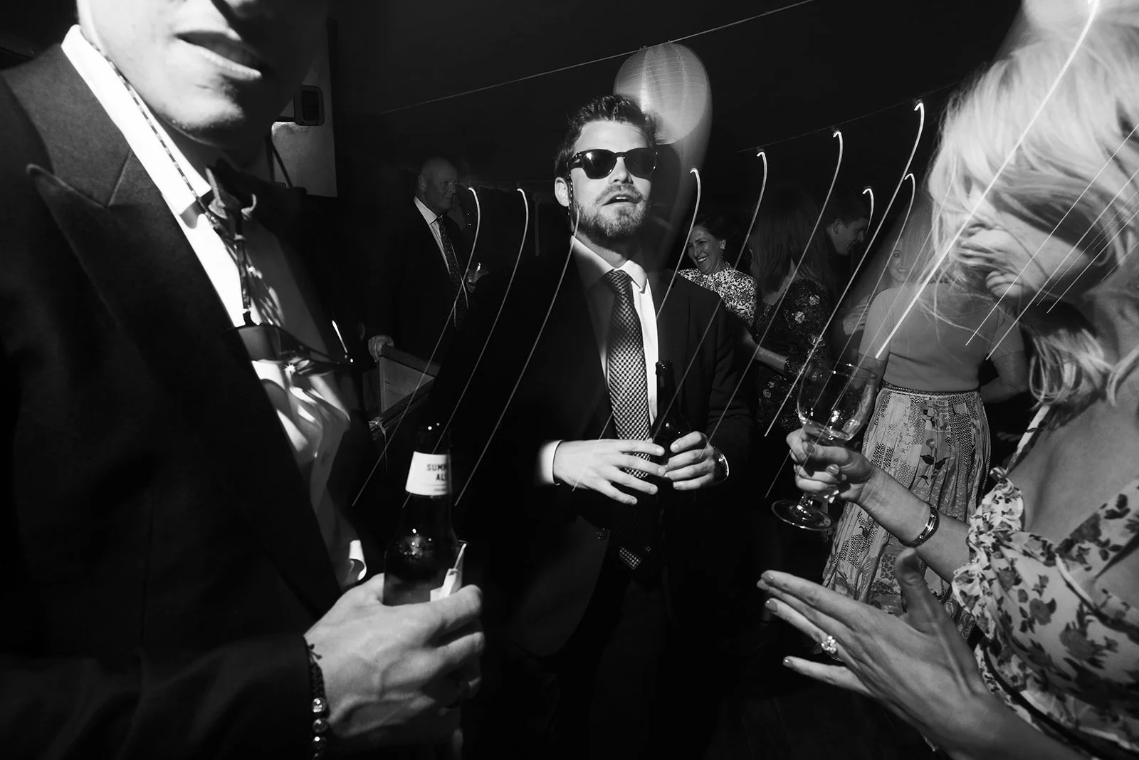People socializing at a party, holding drinks, some wearing sunglasses, in a dimly lit setting.