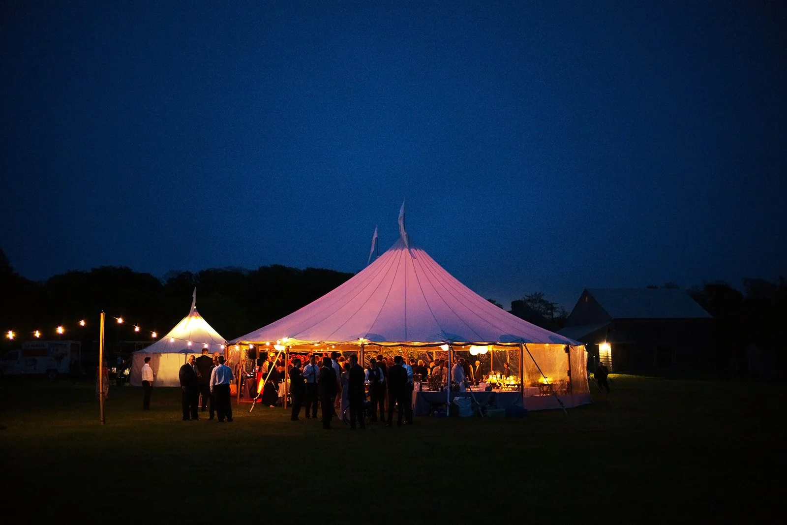 A large event tent illuminated at night with people gathered underneath. The tent has a pointed top and is set on a grassy field with string lights and a smaller tent nearby.