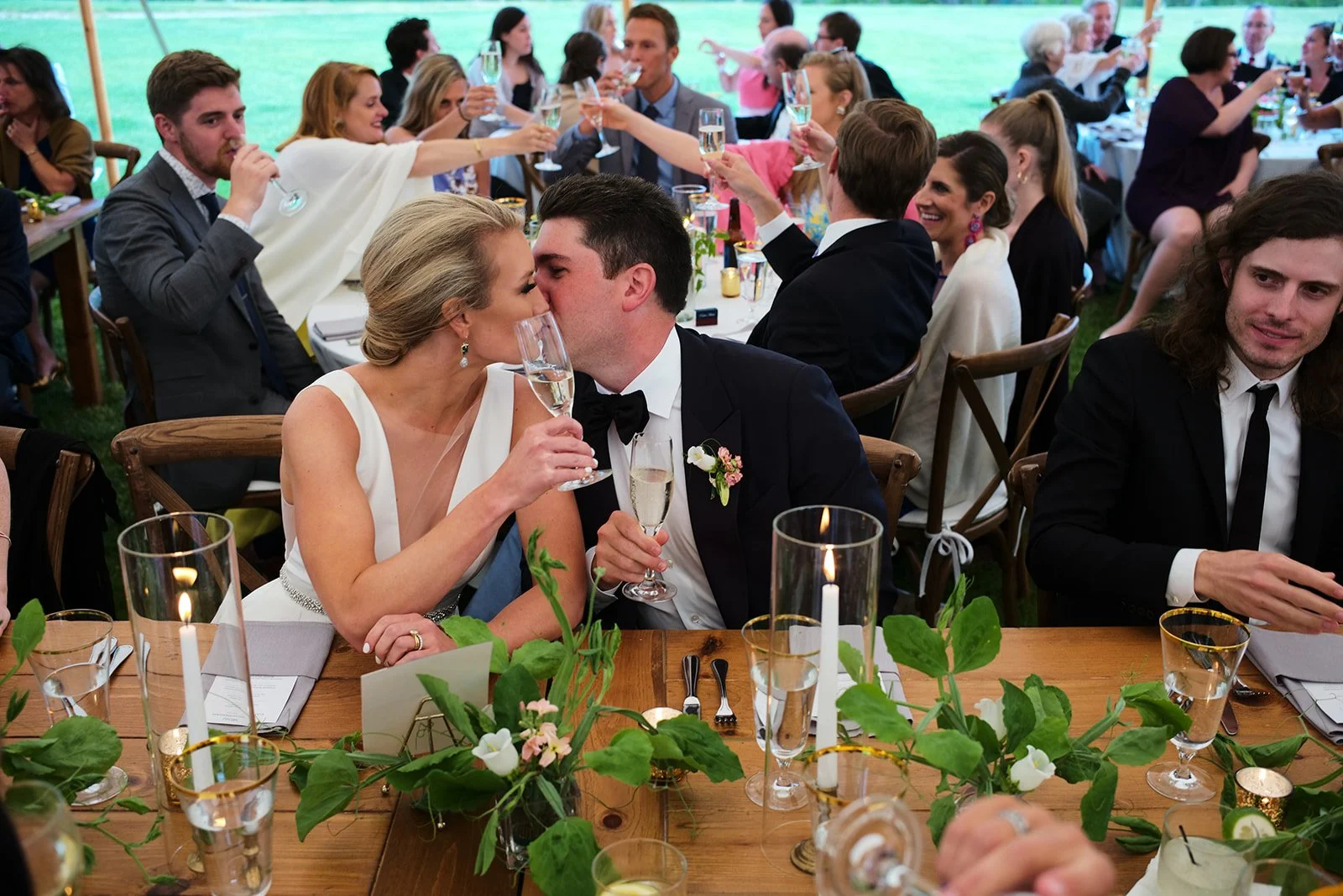 Couple sharing a kiss at a wedding reception, surrounded by seated guests raising glasses for a toast, decorated with flowers and candles.