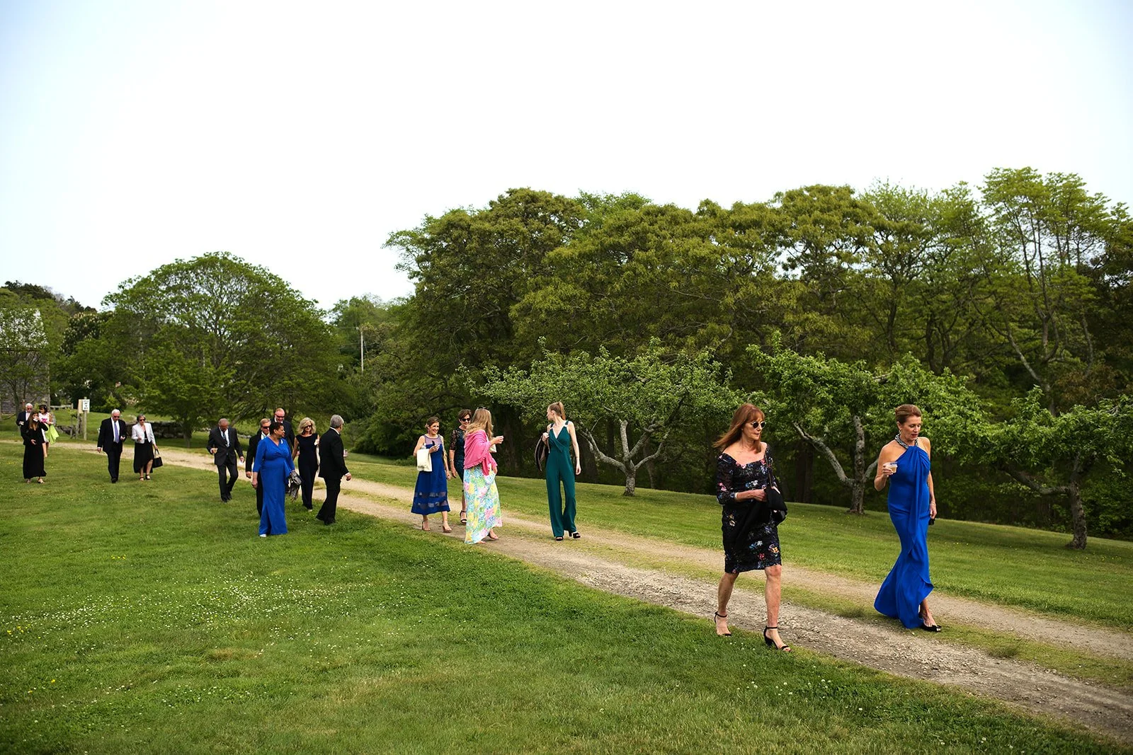 Group of people walking along a gravel path in a lush green park, dressed in formal attire, with trees and a grassy area surrounding them.