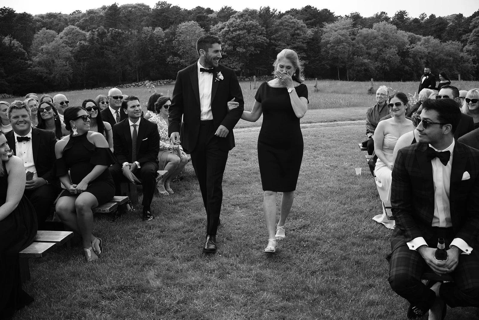 A wedding ceremony outdoors with guests seated on benches. A man in a tuxedo and a woman in a black dress walk arm-in-arm, the woman covering her mouth with her hand. The guests are smiling and watching the couple.