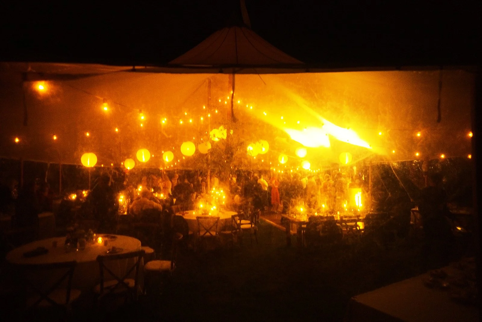 Nighttime scene of a decorated outdoor party under a tent with yellow hanging lanterns and string lights, with people gathered around tables.