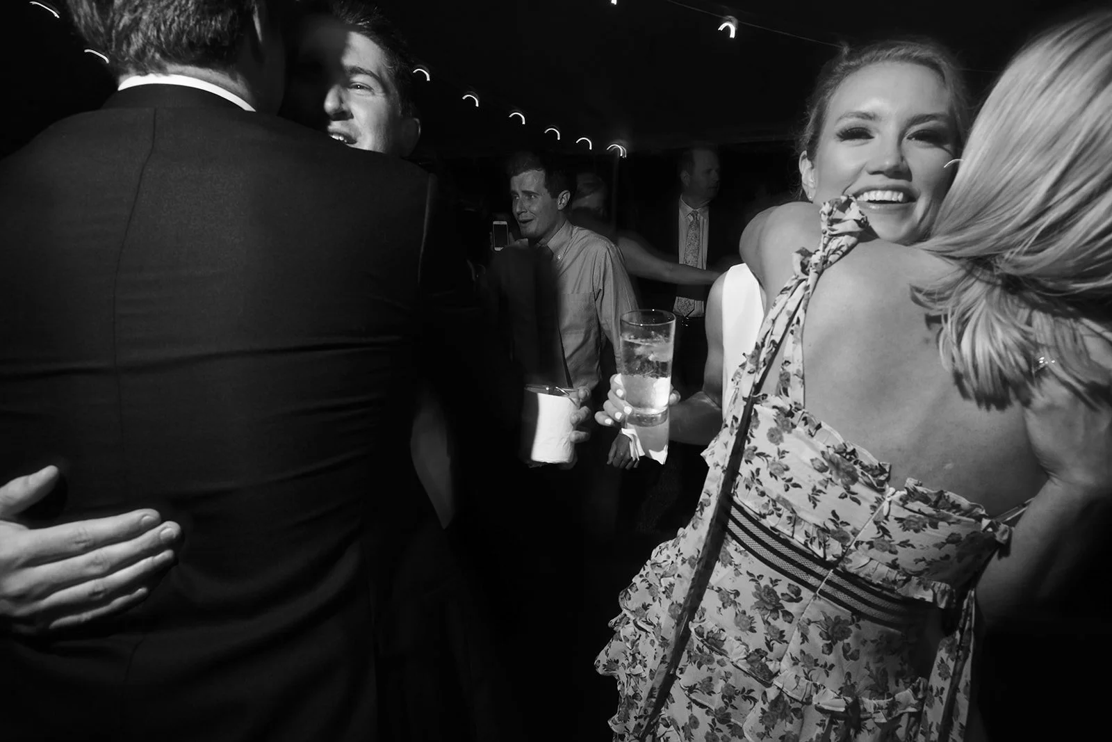 People dancing and hugging at a party, some holding drinks, with smiling faces, in a crowded indoor setting. Black and white photo.
