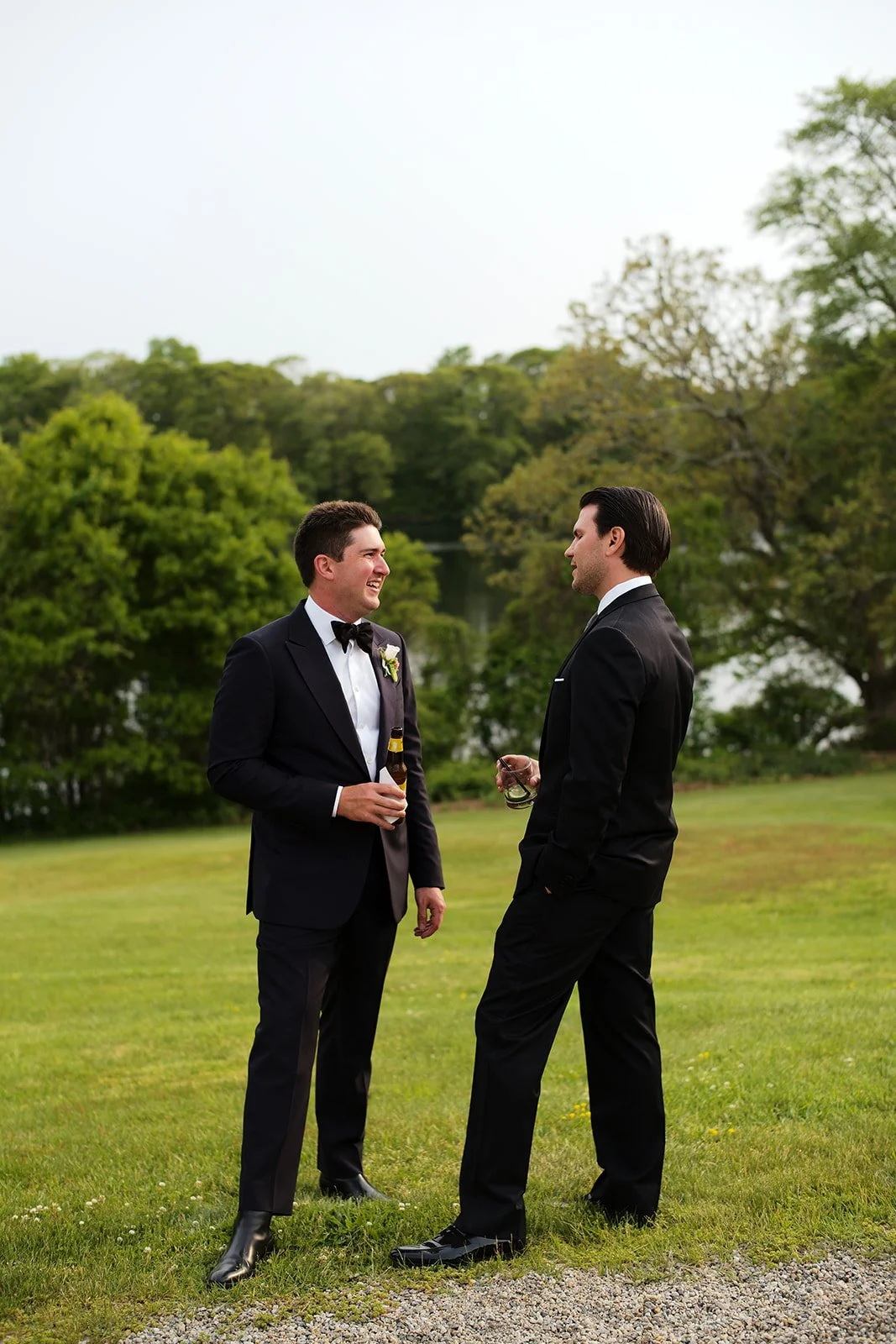 Two men in tuxedos standing outdoors, smiling and conversing, one holding a beer and the other a glass of drink, with greenery and trees in the background.