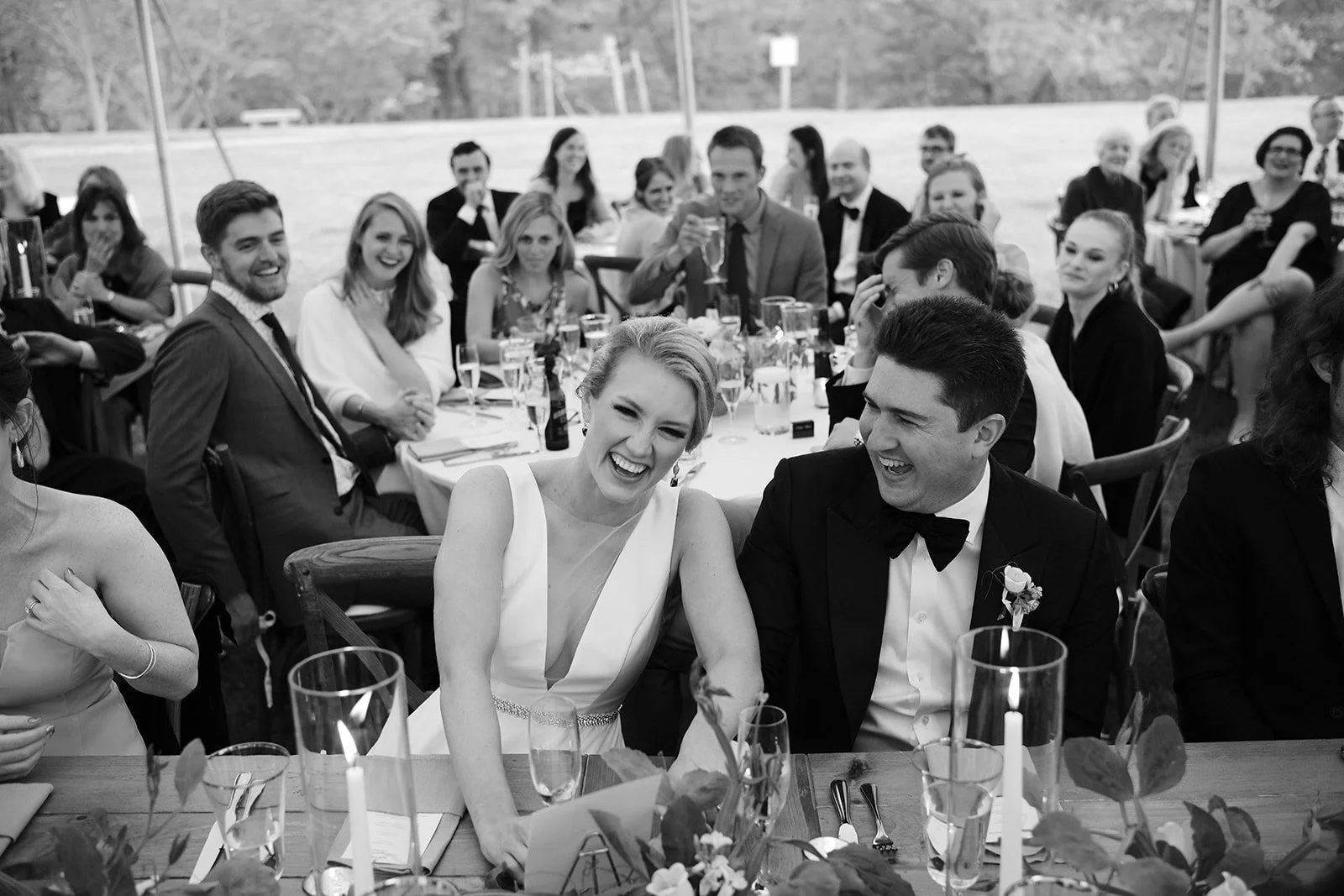 A group of people at a wedding reception, including a bride and groom at the center, smiling and laughing during the celebration.