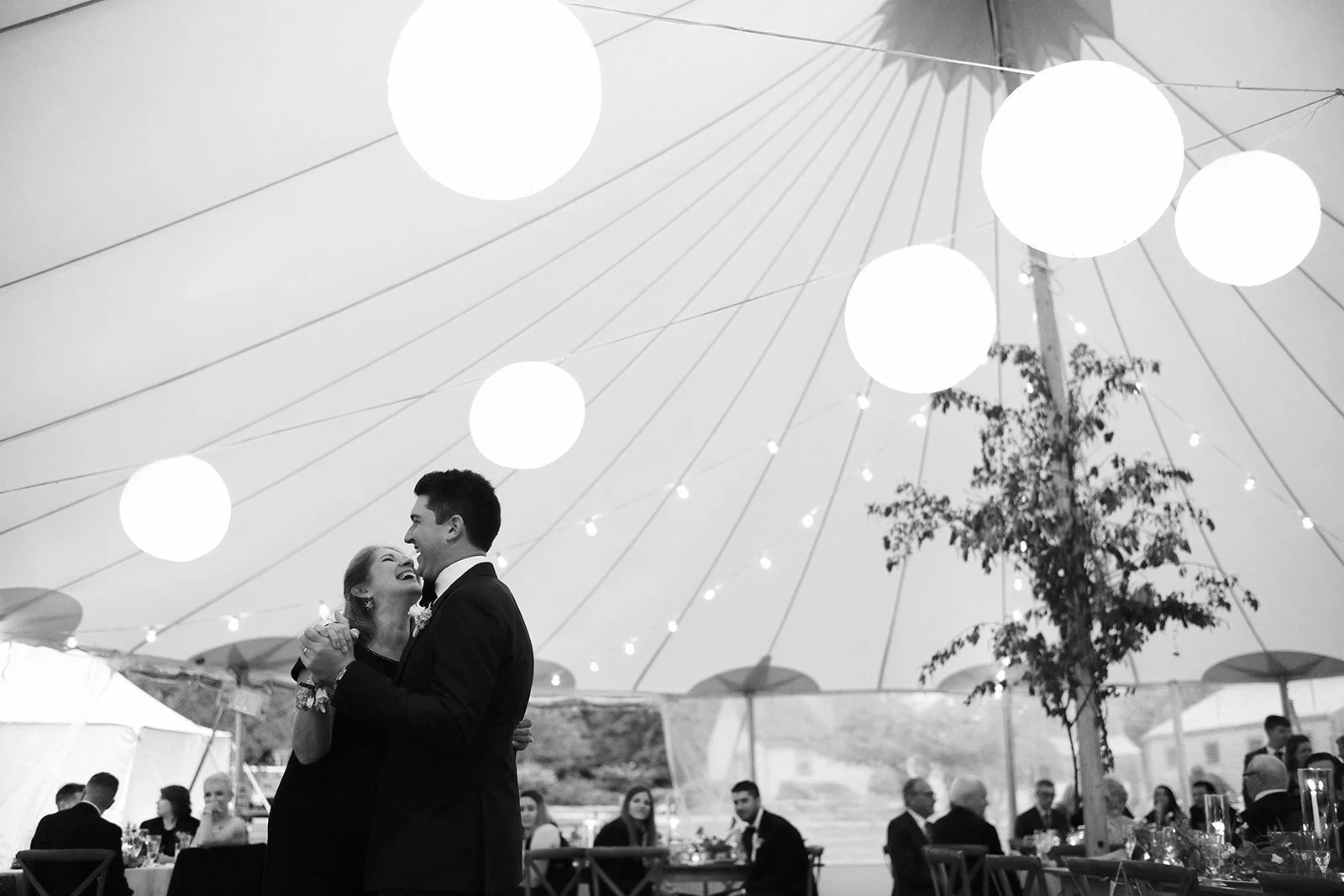 A couple dancing at a wedding reception under a large, decorated canopy with hanging lights and lanterns, with seated guests in the background.