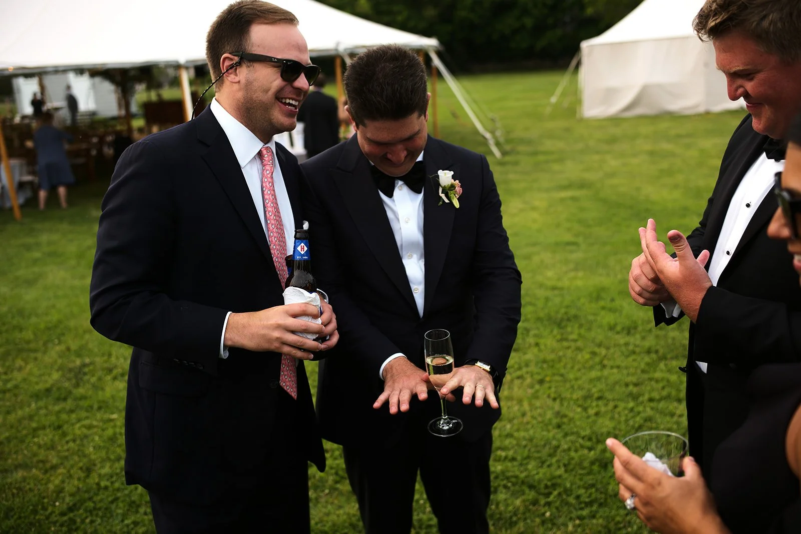 Men in tuxedos at an outdoor wedding celebration, raising drinks and smiling.