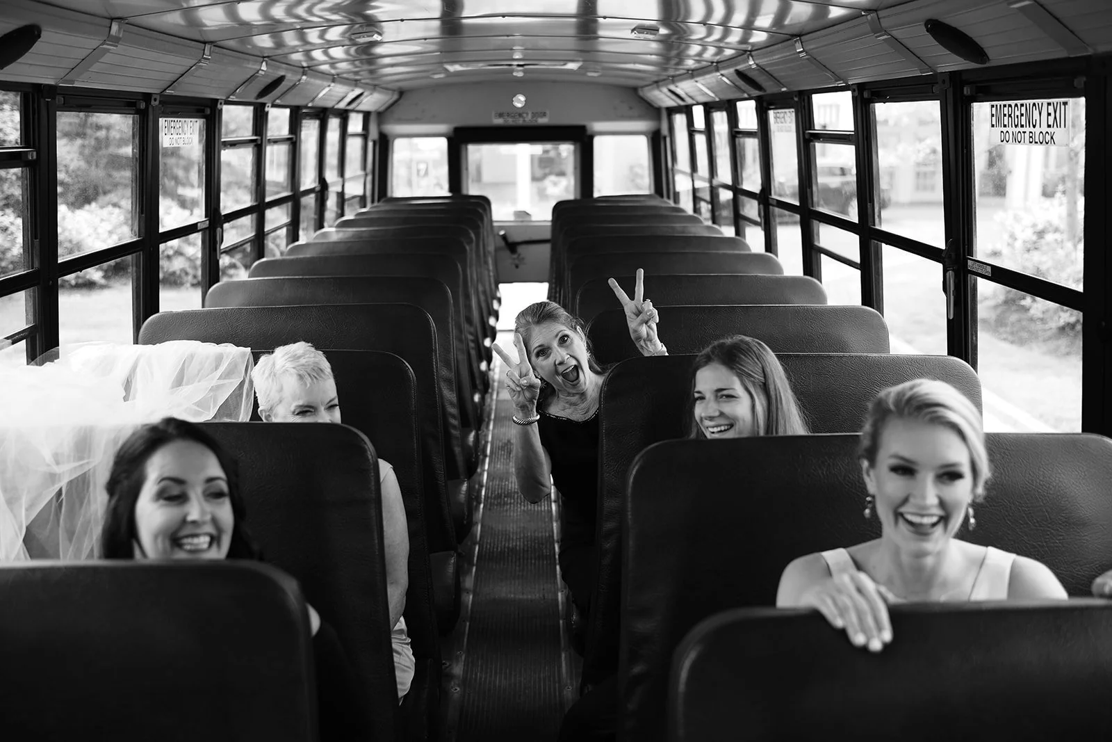 Five women sitting on a school bus, with one woman in the middle making peace signs and all smiling.
