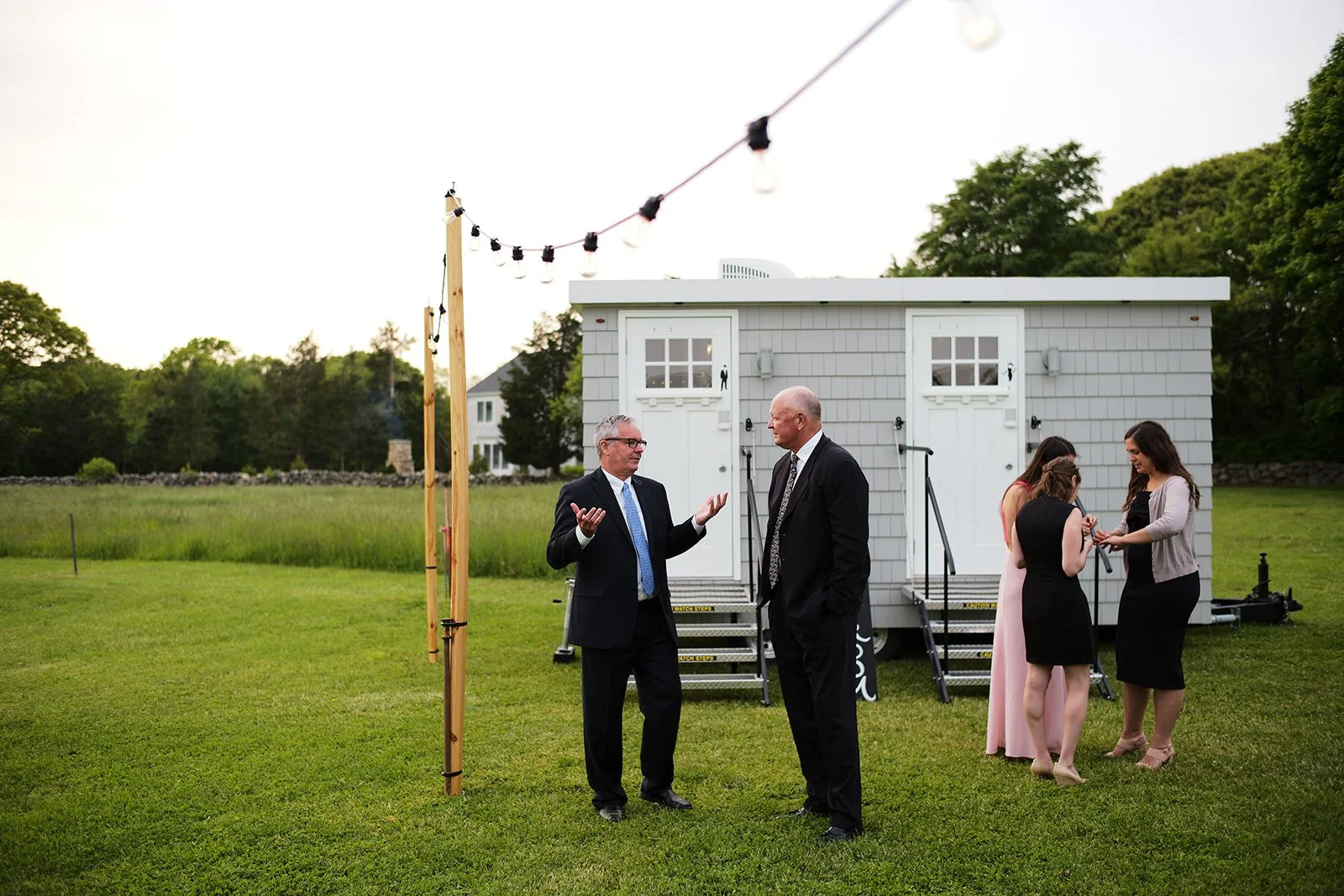 Four people, two men in suits and two women in dresses, standing outside on a grassy area near a small white building with two doors. One man is speaking, gesturing with his hands, while the others listen. In the background, there are trees and a lar