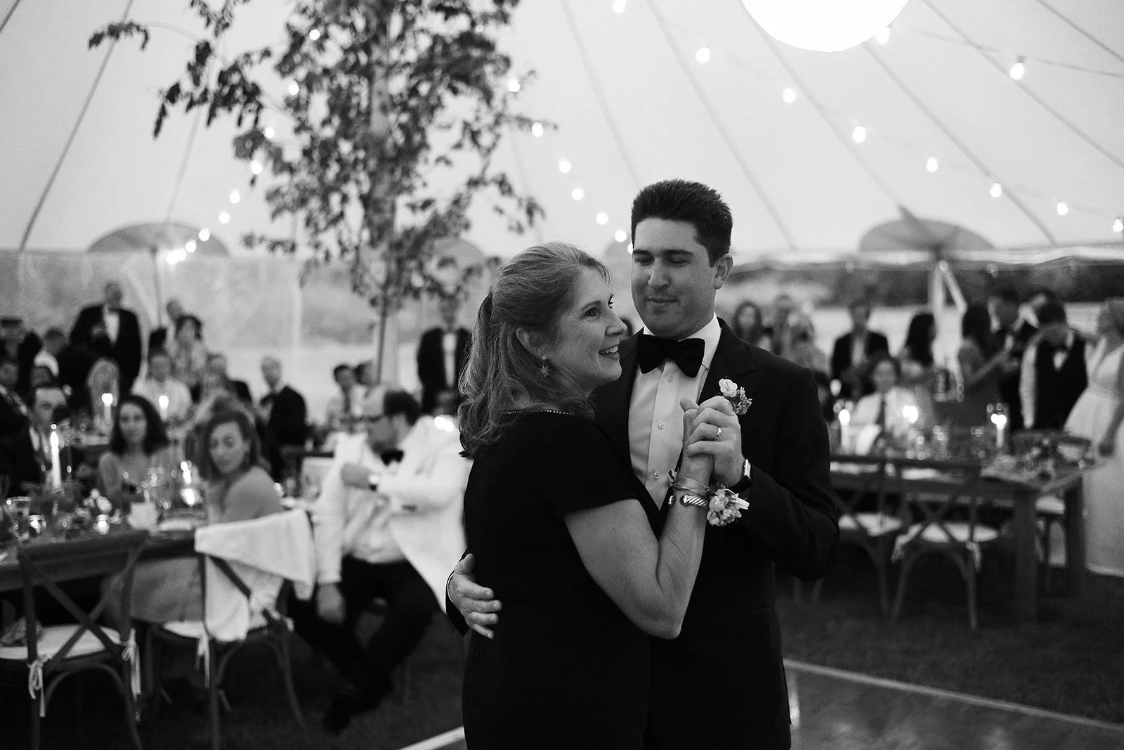 A mother and son dance at a wedding reception under a large tent, surrounded by seated guests, string lights, and a small tree in the background.