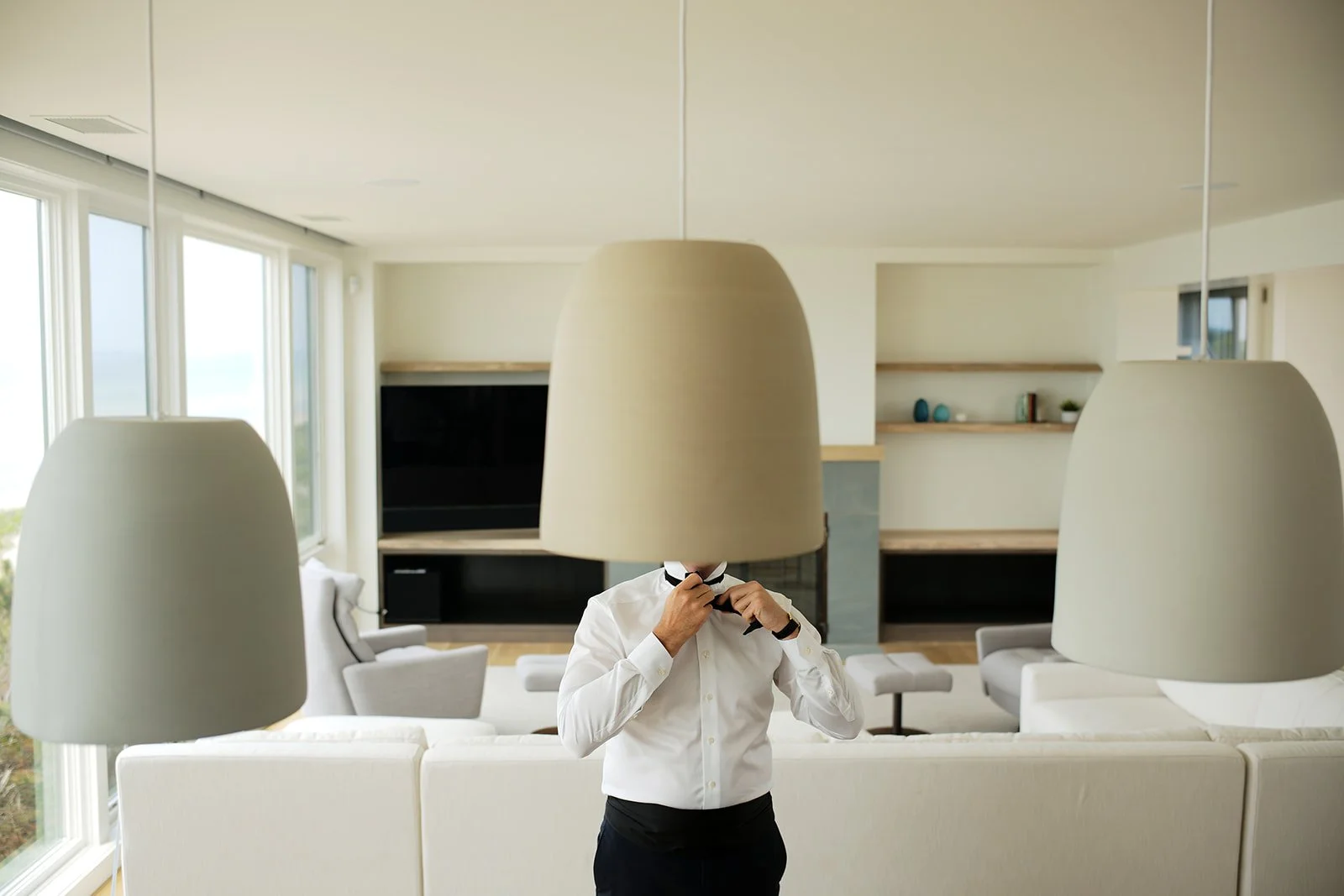 Person adjusting a black tie while standing in a modern, well-lit living room with large windows, beige sofa, and hanging ceiling lamps.