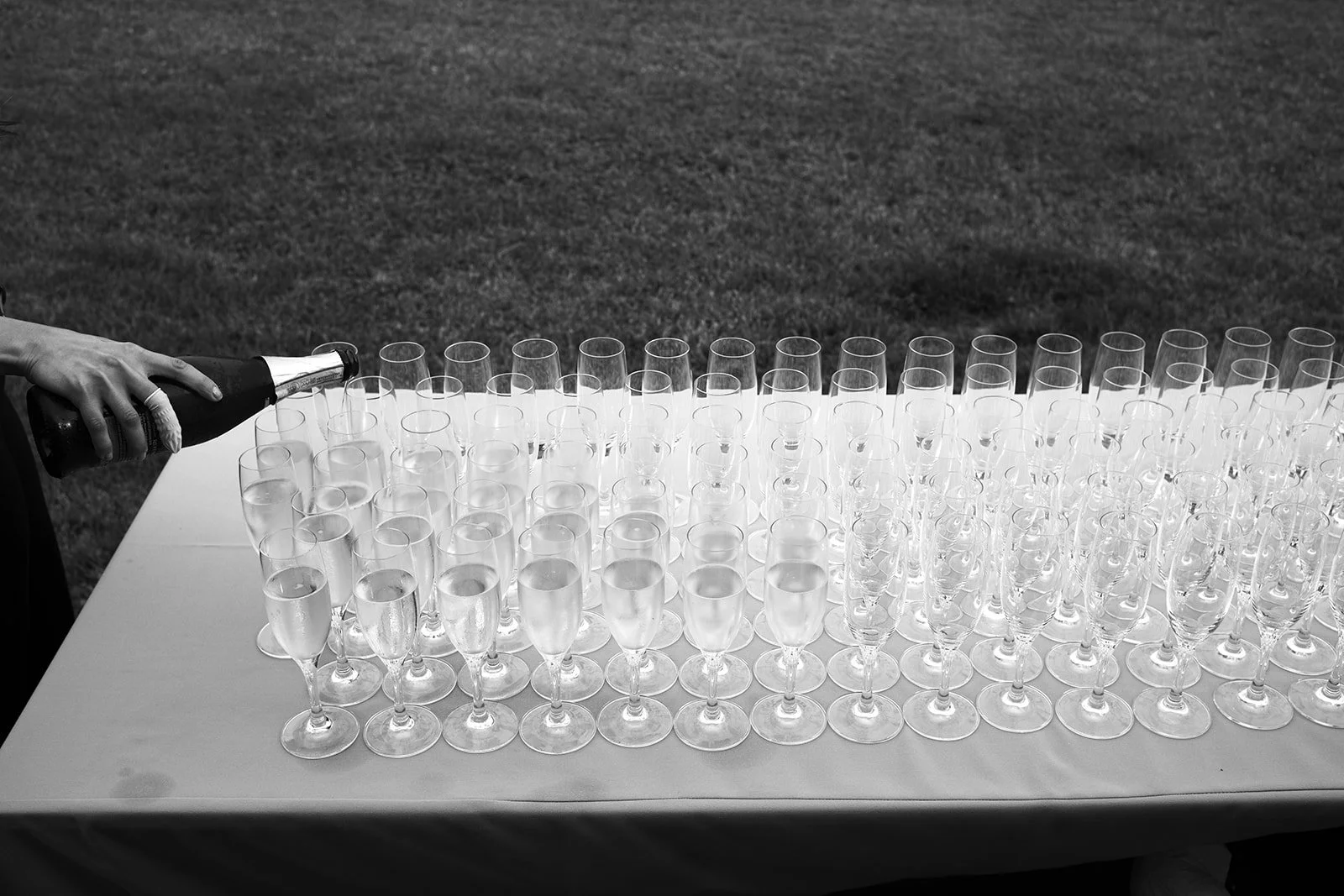 Person pouring champagne into a row of empty champagne glasses on a table outdoors.