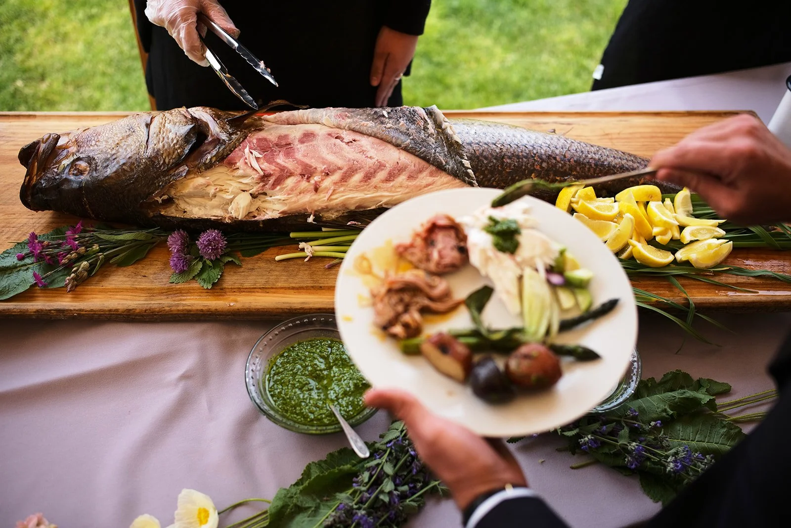 People serving and preparing a whole fish at a buffet or event, with lemon slices, herbs, and a green sauce on a wooden table.