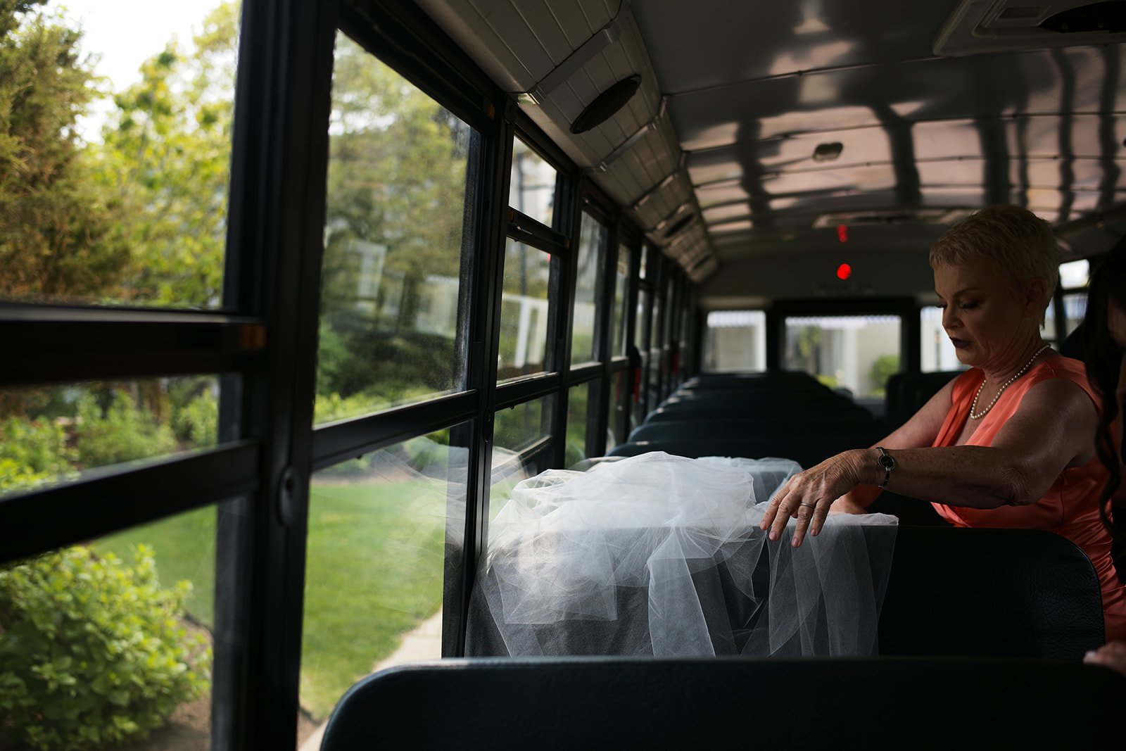 An older woman with short blonde hair, wearing a peach sleeveless dress, sitting inside a bus with large windows, adjusting a white veil or fabric on her lap, with a green outdoor scene visible through the windows.