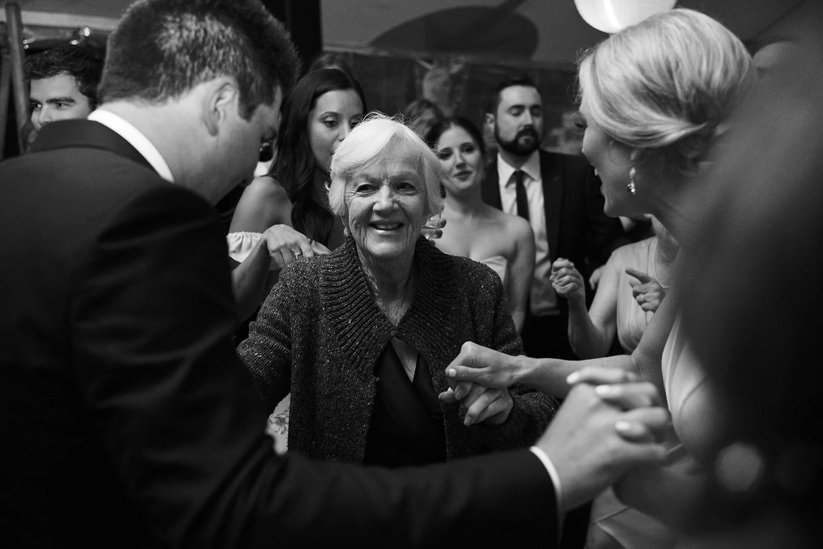 An elderly woman smiling and holding hands with a young man at a social gathering, with several people in the background engaging and talking.