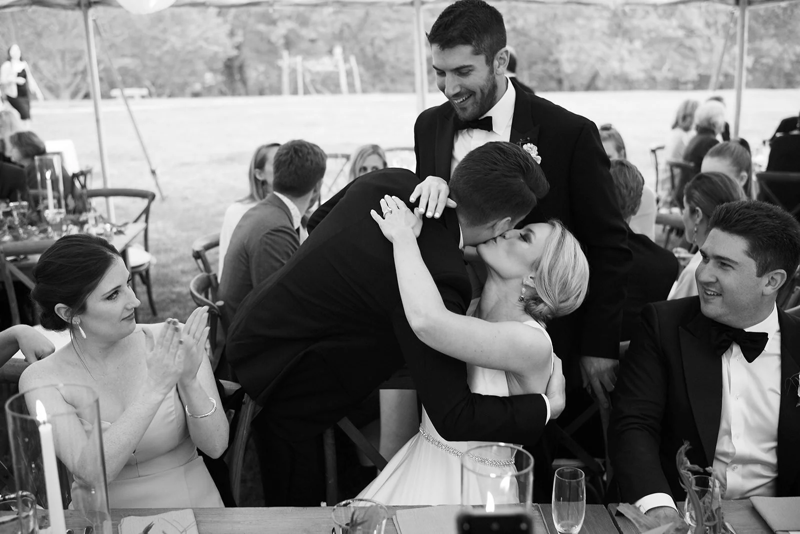 A wedding celebration with a couple kissing and others around them, including a woman clapping, at an outdoor event under a tent.