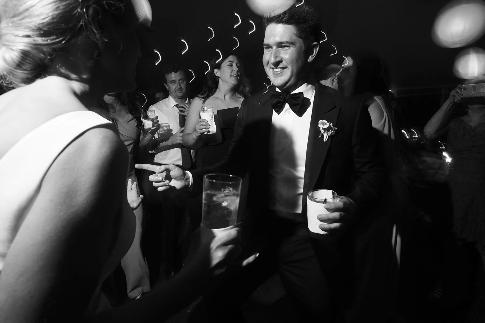Black and white photo of a man in a tuxedo with a bow tie, smiling and holding a drink, at a social gathering or wedding reception with several people around him, some holding drinks and engaging in conversation.