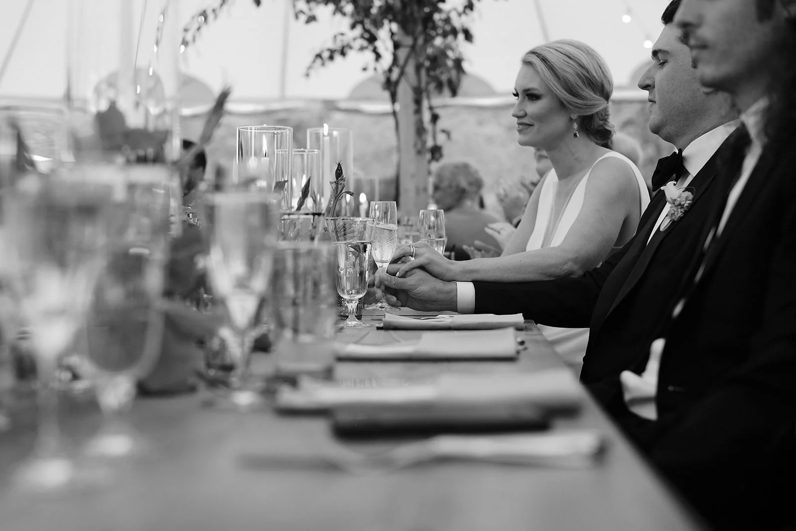 Black and white photograph of a bride and groom holding hands across a banquet table at their wedding reception, with guests and floral decor in the background.