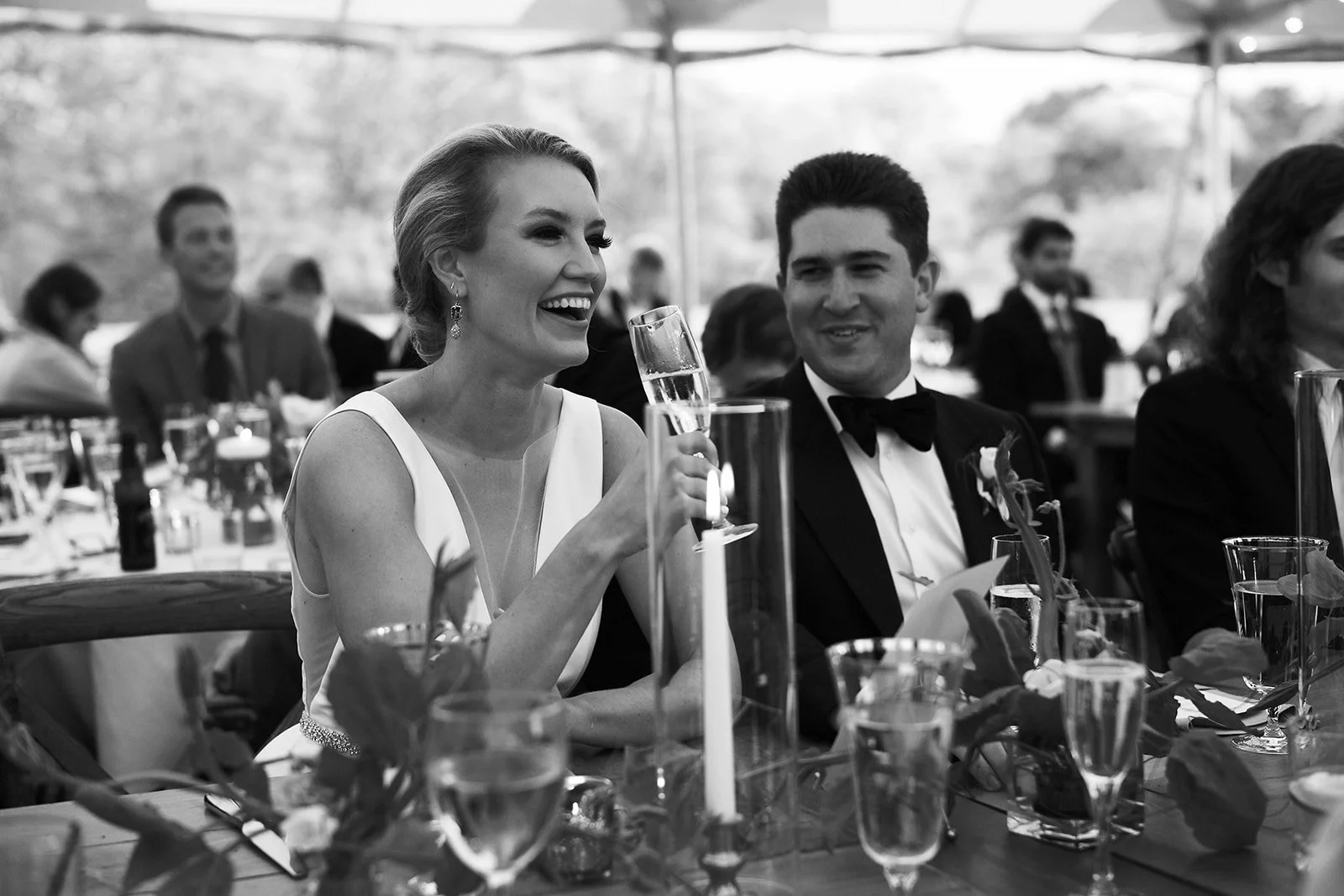 Black and white photo of a smiling woman in a sleeveless dress holding a glass of champagne at a formal event, seated at a decorated table with a man in a tuxedo on her right, in an outdoor setting with other people in the background.
