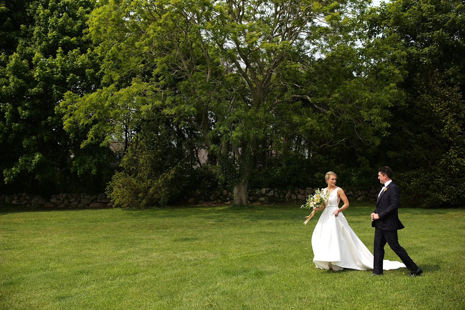 Bride in a white wedding gown holding a bouquet walking with groom in a black suit on a grassy field with trees in the background.