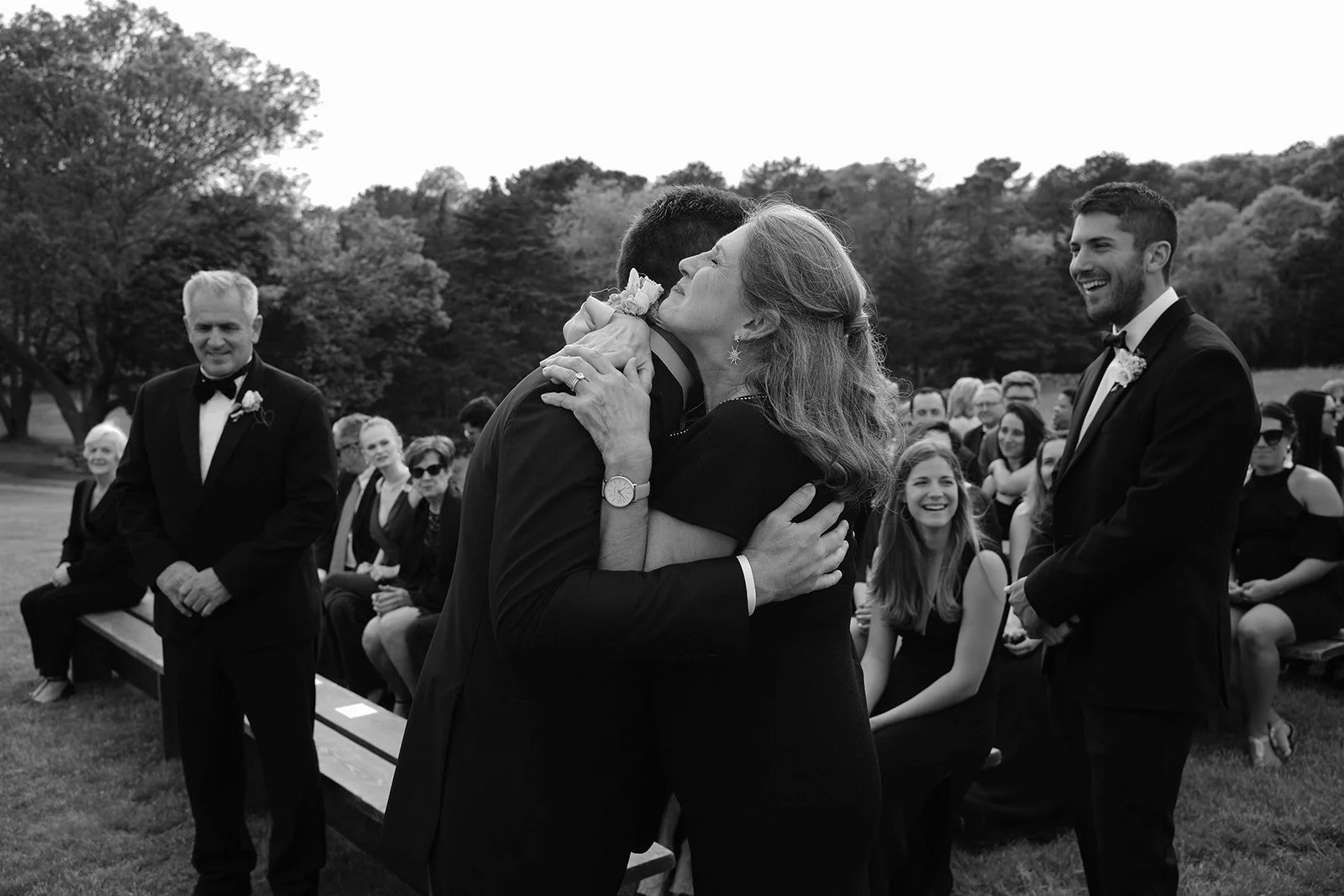 A woman is hugging a man during a wedding ceremony outdoors, with guests sitting behind them and trees in the background.