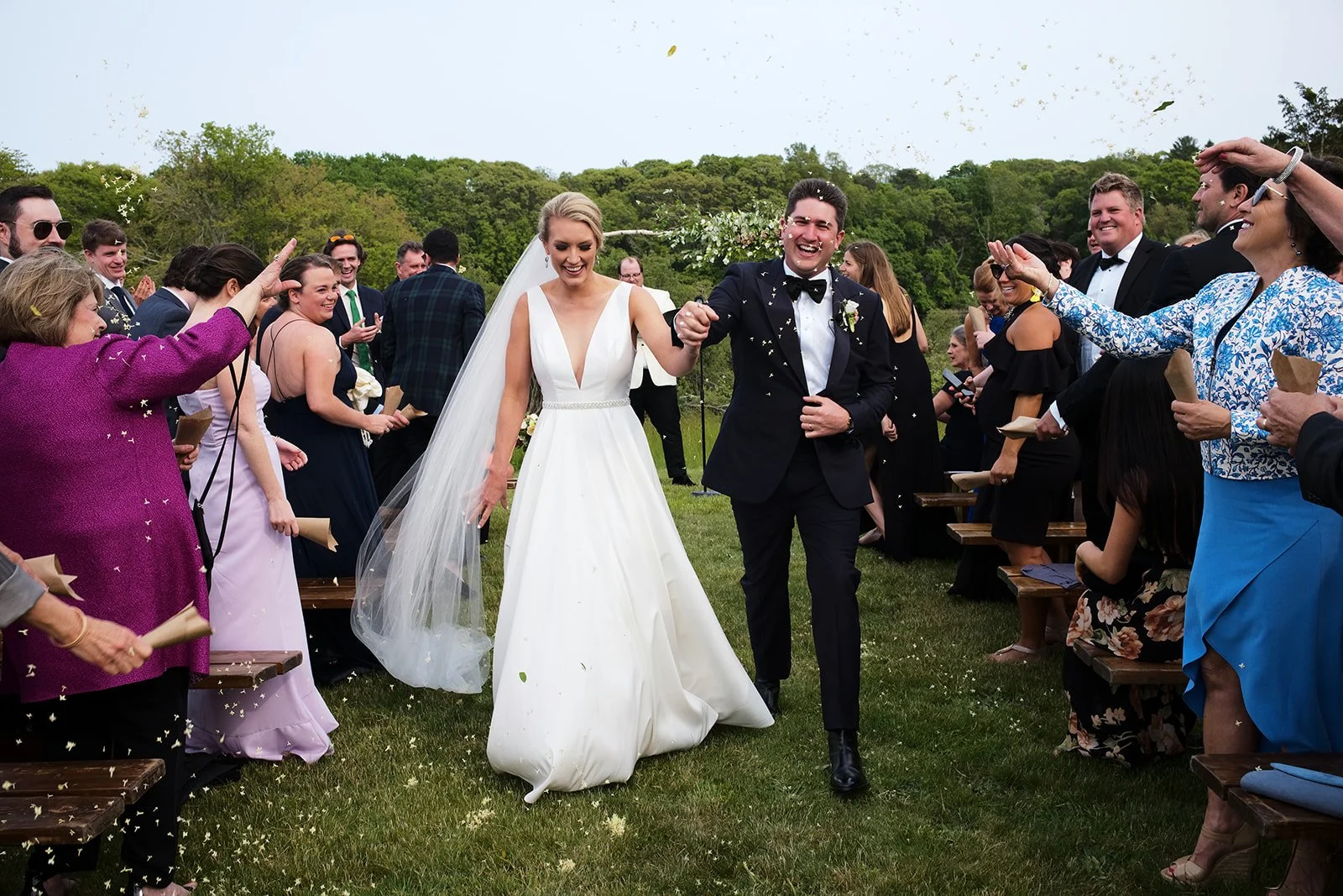 A bride and groom walking arm in arm after their wedding ceremony, surrounded by smiling guests throwing confetti outdoors on a grassy field with trees in the background.
