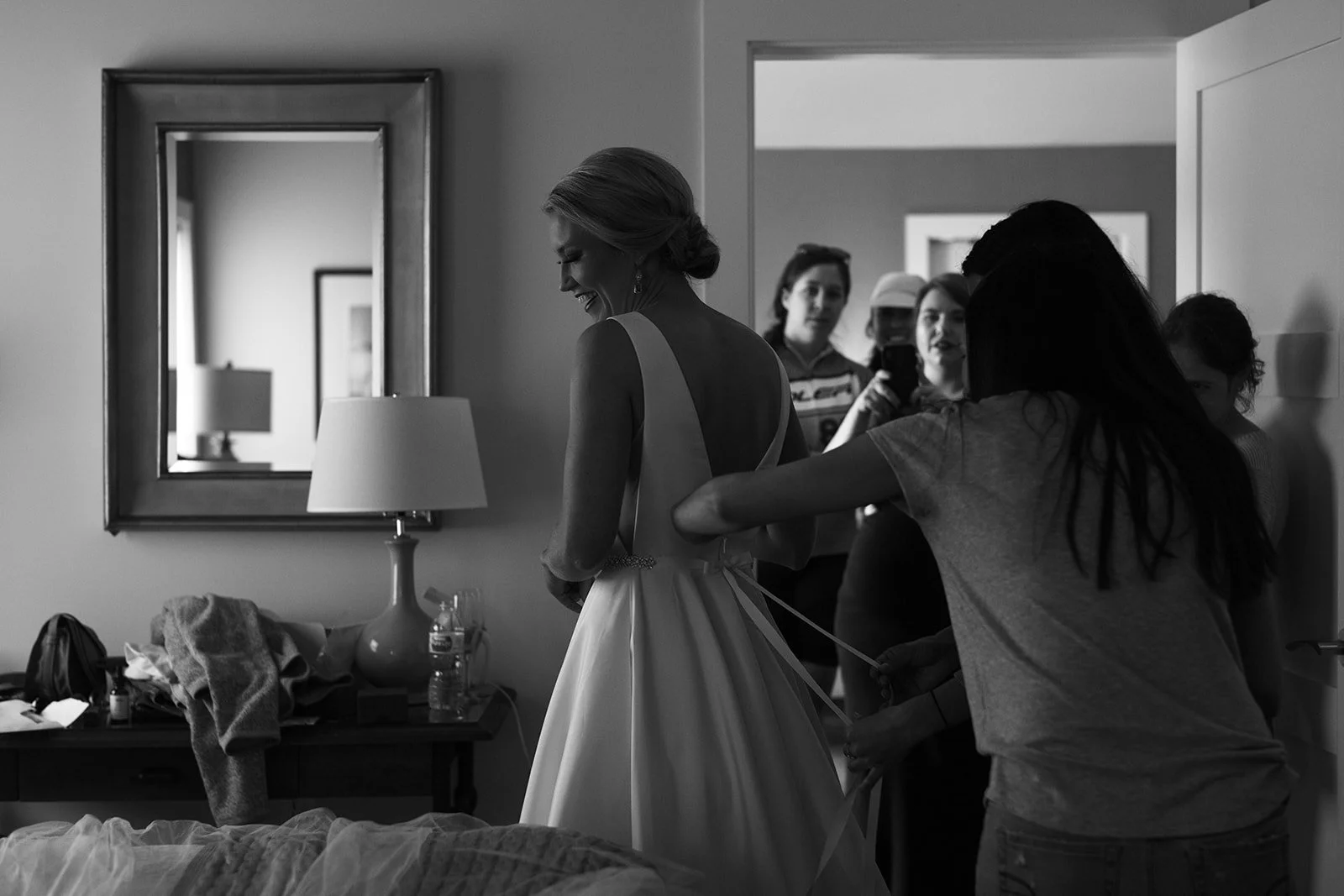 A bride in a wedding dress is getting ready while friends help her, in a room with a mirror and a lamp.