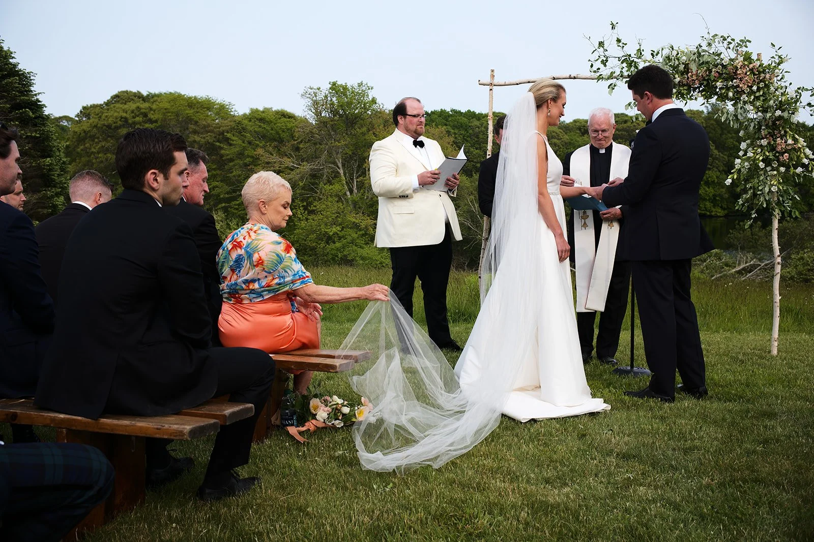 Bride and groom exchanging vows at an outdoor wedding ceremony with guests watching. Green trees and grass surround them, and there is a floral arch behind the couple.
