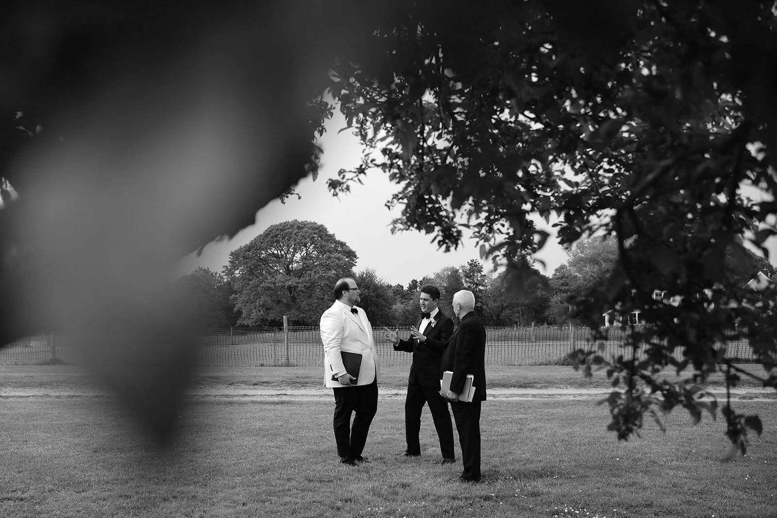 Three men in formal tuxedos having a discussion outdoors, framed by leaves in the foreground, with trees and a fence in the background.