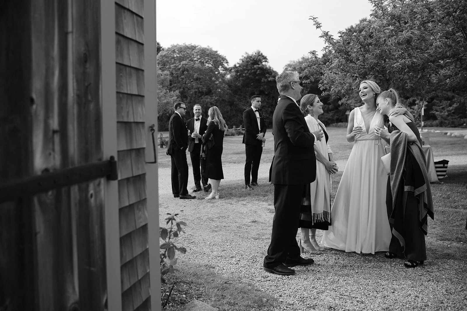 Group of elegantly dressed people socializing outdoors, with some wearing tuxedos and others in wedding attire, in a park-like setting with trees in the background.