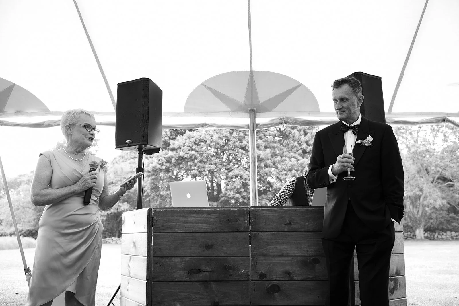 A wedding celebration outside under a tent. An elderly woman is speaking into a microphone, and a man in a tuxedo is holding a glass of champagne, looking at her. A DJ with a laptop is in the background.
