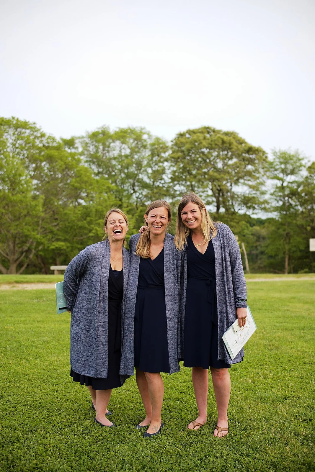 Three women wearing matching navy dresses and gray cardigans smiling and standing together on a grassy area outdoors, with trees and a cloudy sky in the background.