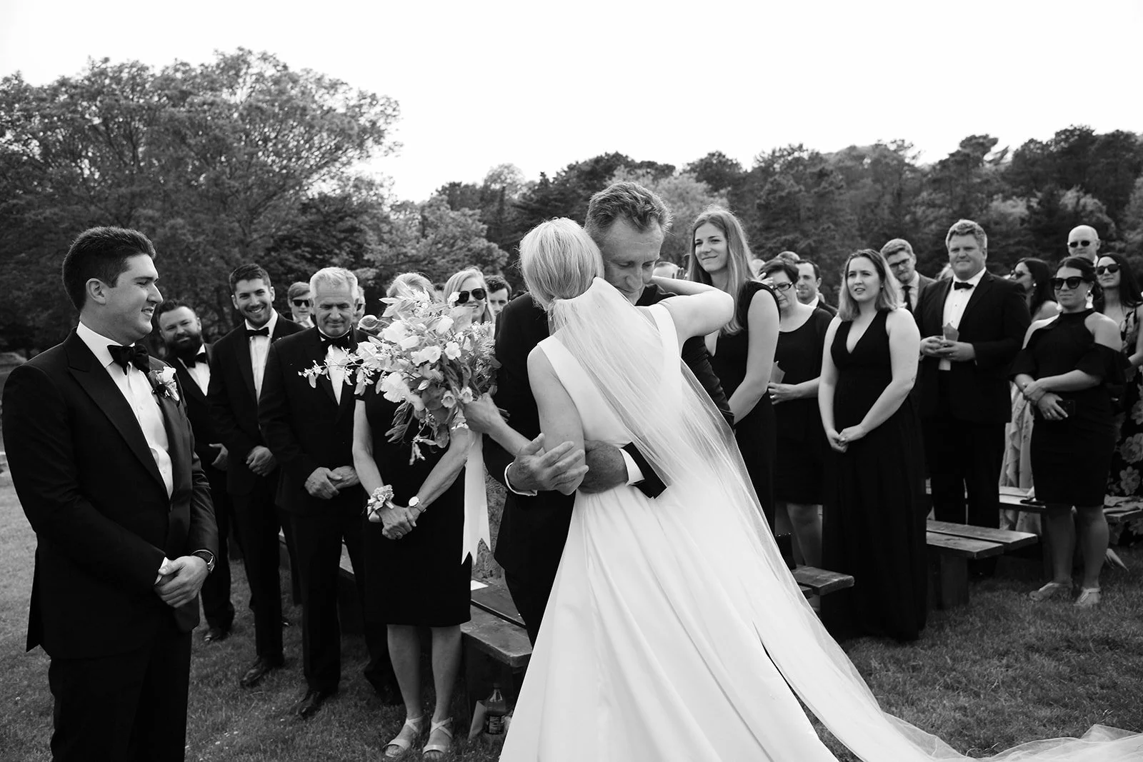 A bride and groom share a dance at their outdoor wedding ceremony, surrounded by guests dressed in formal attire.