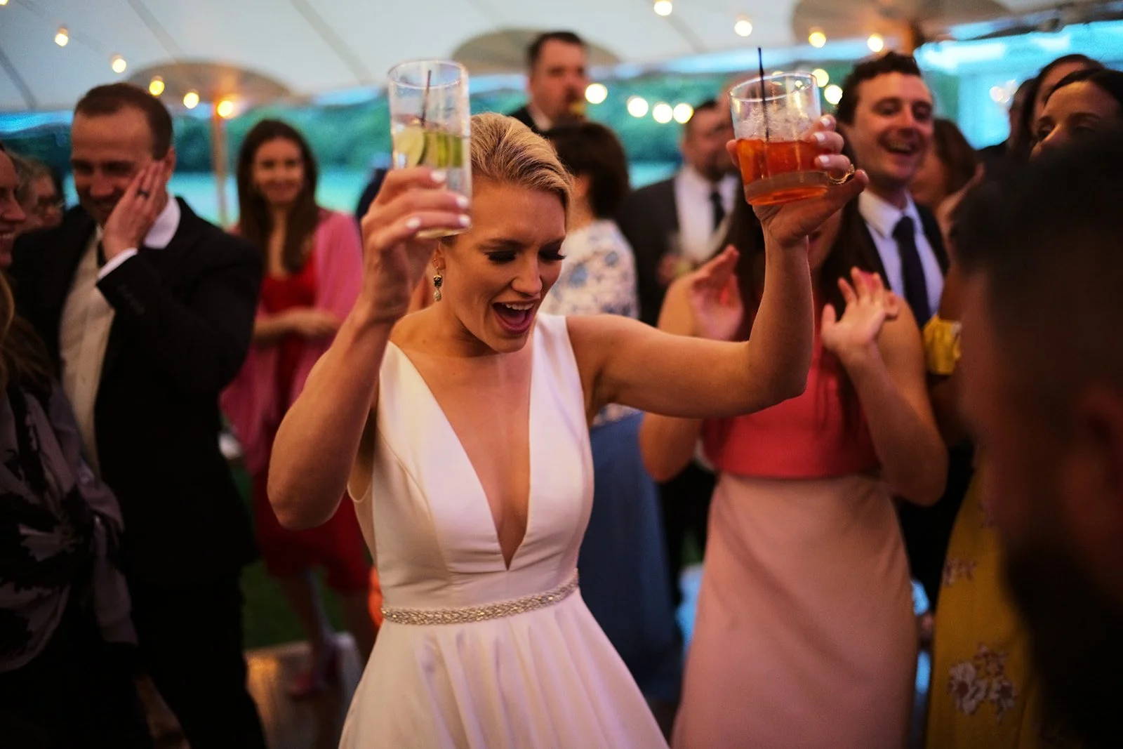 A woman in a white dress dancing and celebrating at a party with people around her, holding drinks in a festive atmosphere under a tent with string lights.