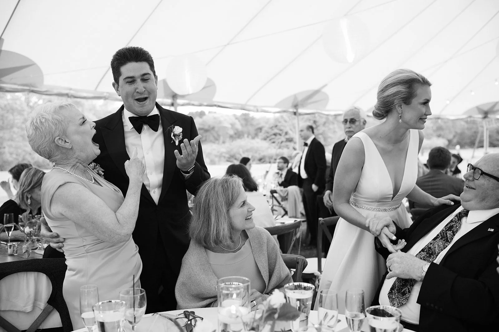 Black and white photo of a wedding reception with people talking and smiling, under a canopy. Elderly woman and a young man in a tuxedo are in the foreground, with women sitting and standing nearby.
